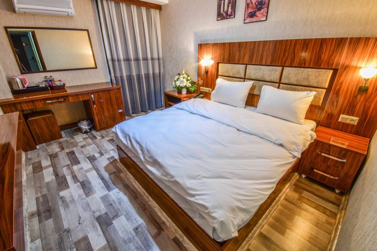 A hotel room with a wooden headboard, a large bed with white bedding, a bedside table with a lamp, a mirror, and a small desk with a kettle and a flower arrangement.