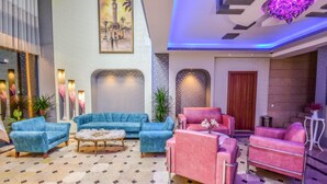 A lobby with a blue sofa set, a pink sofa set, and a wooden coffee table. There is a painting of a building on the wall, and a door is visible in the background. - Marina Hotel (Izmir)