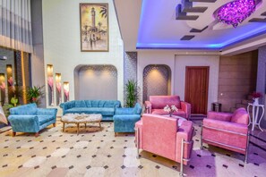 A lobby with a blue sofa set, a pink sofa set, and a wooden coffee table. There is a painting of a building on the wall, and a door is visible in the background. - Marina Hotel (Izmir)