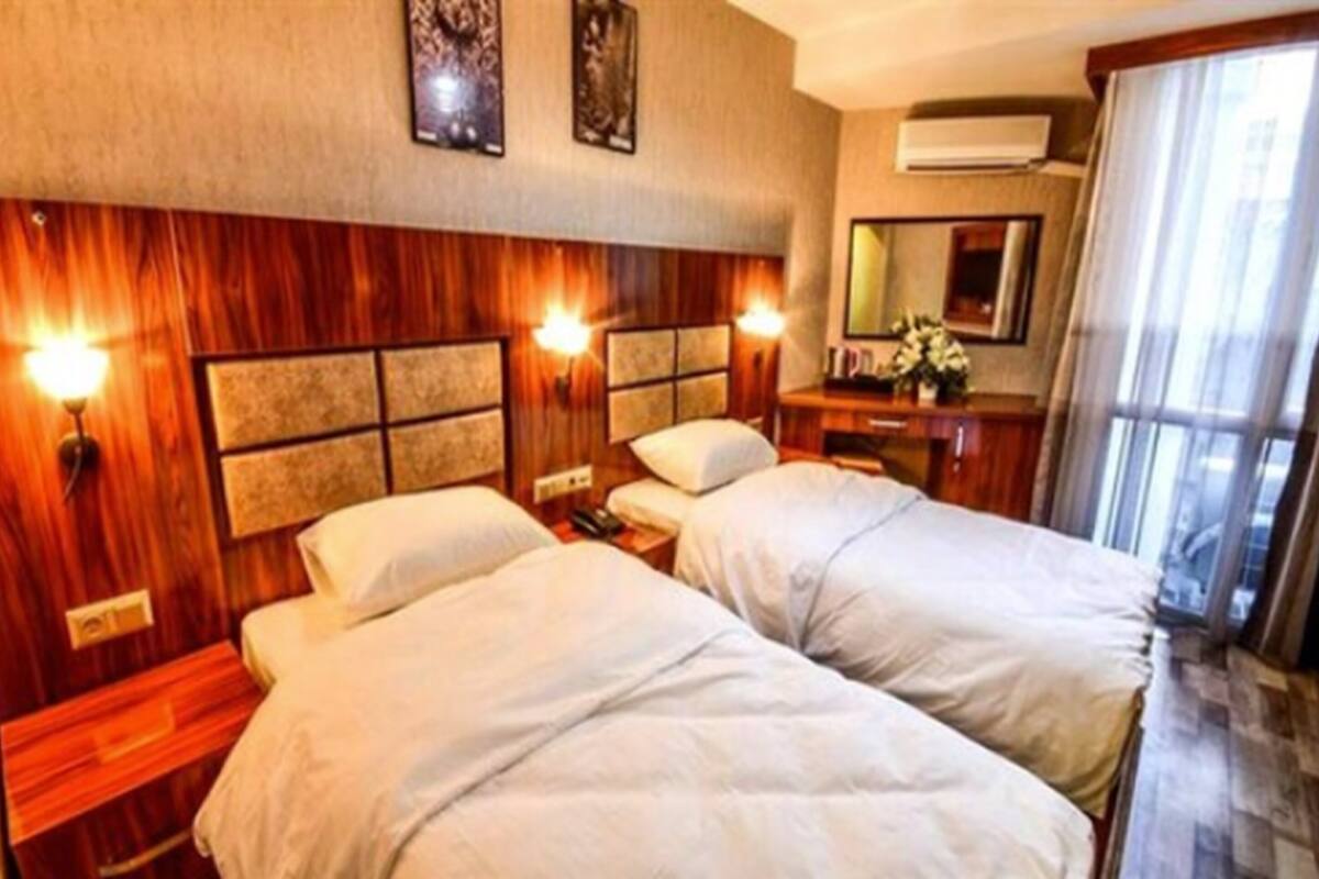 A hotel room with two beds, a wooden headboard, wall-mounted lights, a mirror, and a window with curtains.