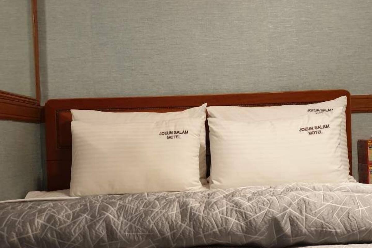 Ein Hotelzimmer mit einem Bett, ausgestattet mit einer strukturierten, grauen Tagesdecke und zwei Kissen mit dem Namen des Hotels.