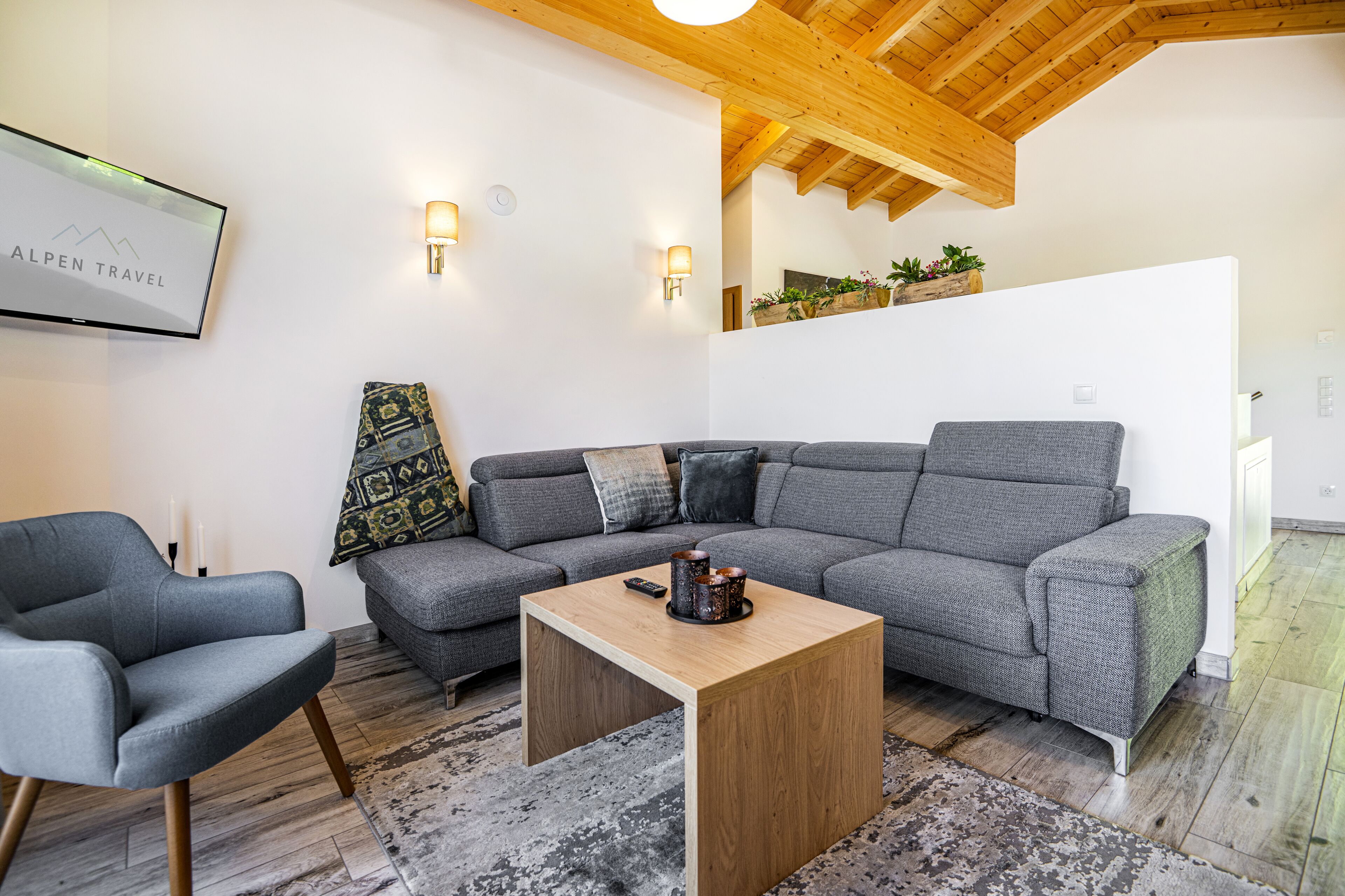 Uma sala de estar moderna com um sofá seccional cinza, uma mesa de centro de madeira e uma poltrona cinza. Há uma imagem emoldurada na parede e uma pequena planta em uma estante.