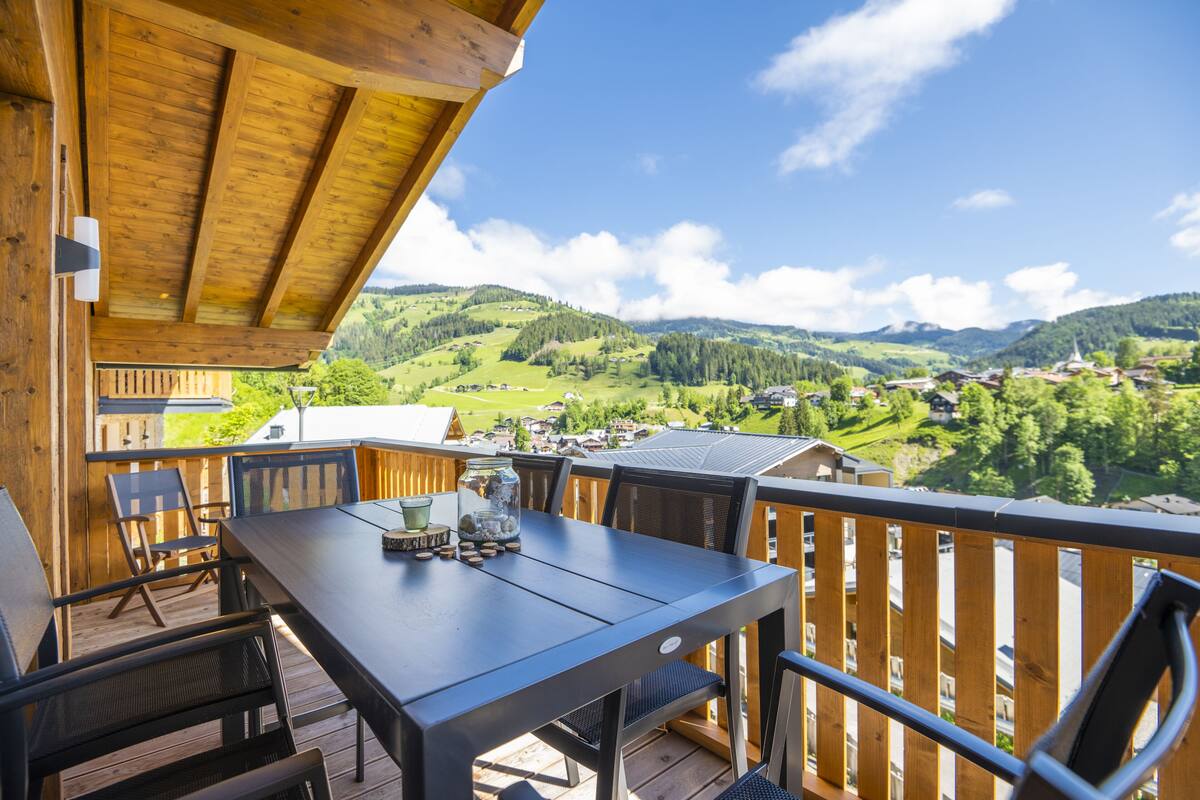 Een houten terras met een tafel en stoelen, met uitzicht op een groene vallei met huizen en heuvels.