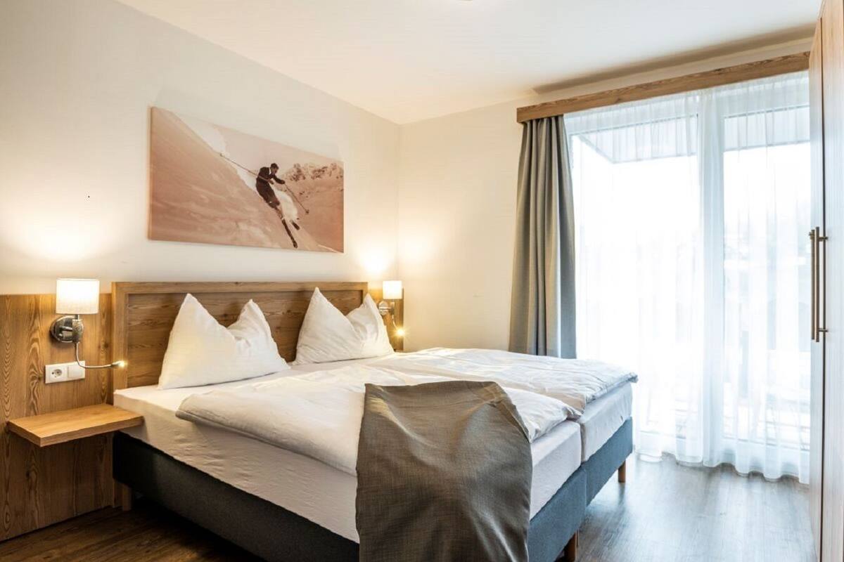 Een hotelkamer met een groot bed, een houten hoofdeinde, twee nachtkastjes met lampen, een groot raam met gordijnen en een schilderij aan de muur.