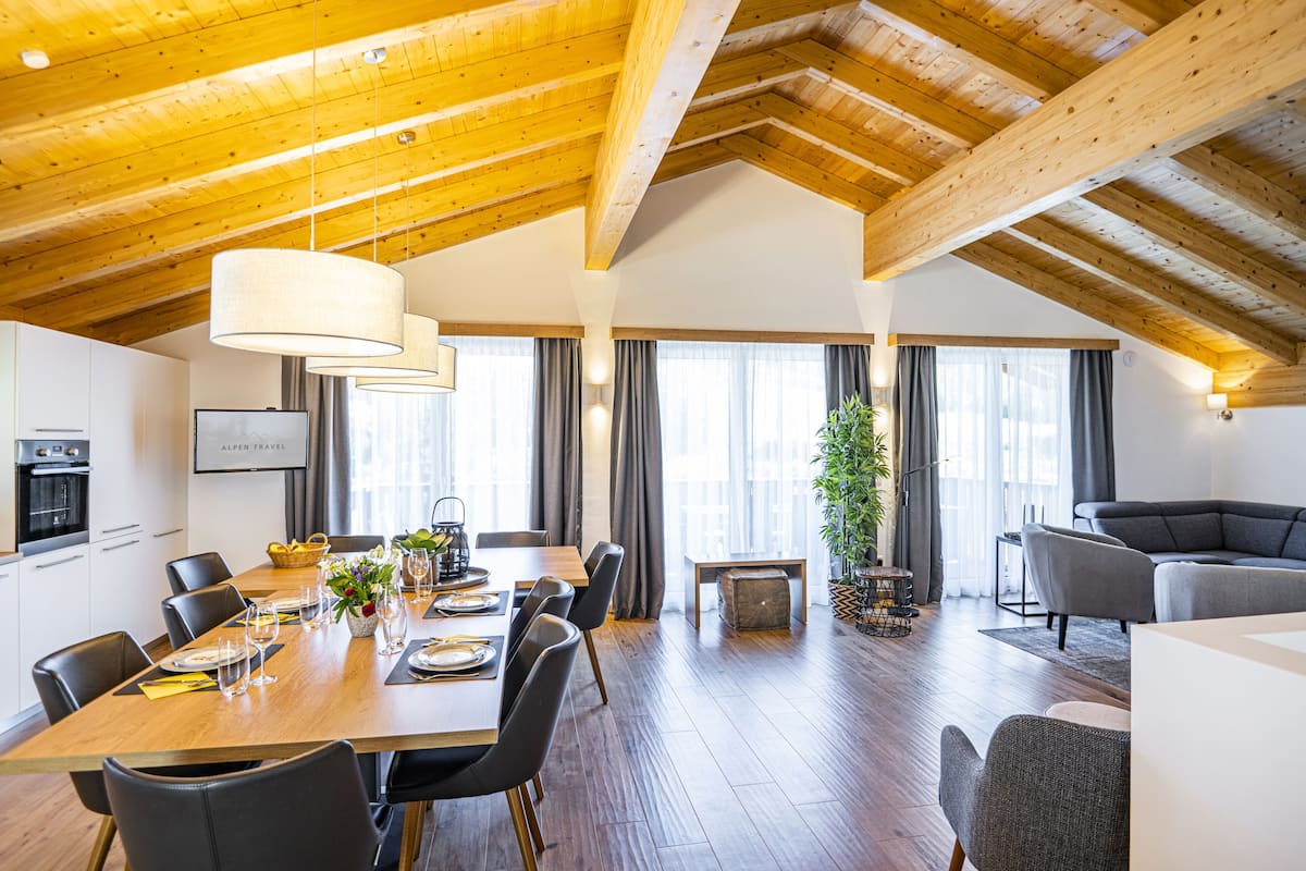 Een ruime eethoek met een houten tafel voor zes personen, omringd door stoelen. Achtergrond toont een grote plant en een bank.