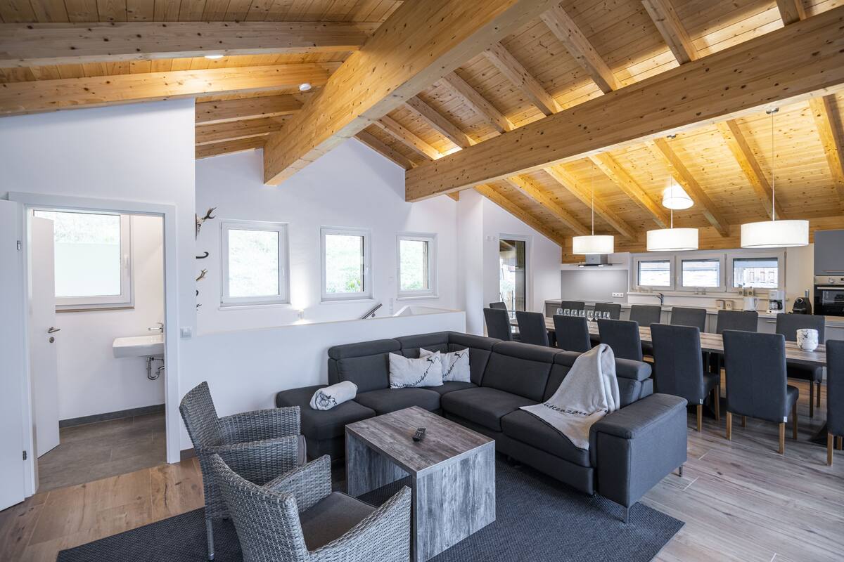 Een moderne woonkamer met een grote hoekbank, een salontafel en rieten fauteuils. Achtergrond toont een keuken met een eettafel en stoelen.