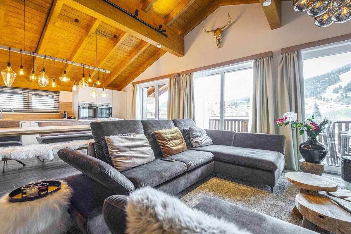 Een moderne woonkamer met een grijze modulaire bank, een houten plafond en een groot raam met uitzicht op een besneeerd landschap.