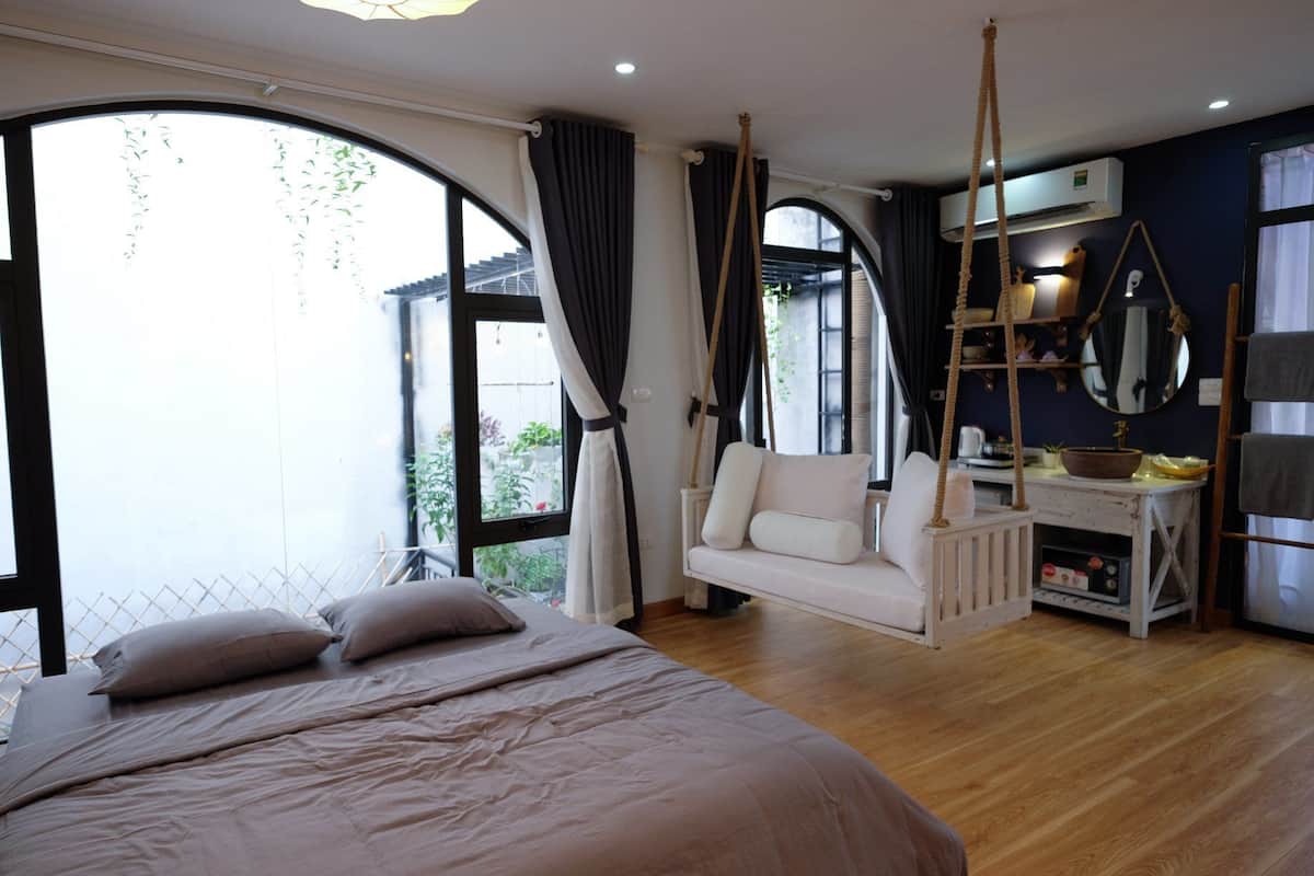 A bedroom with a large bed, a hanging chair, a desk with a mirror, and a view of the outdoors.