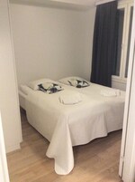 Una cama sencilla e impecable con ropa de cama blanca y dos almohadas, una ventana con cortinas oscuras y suelo de madera.