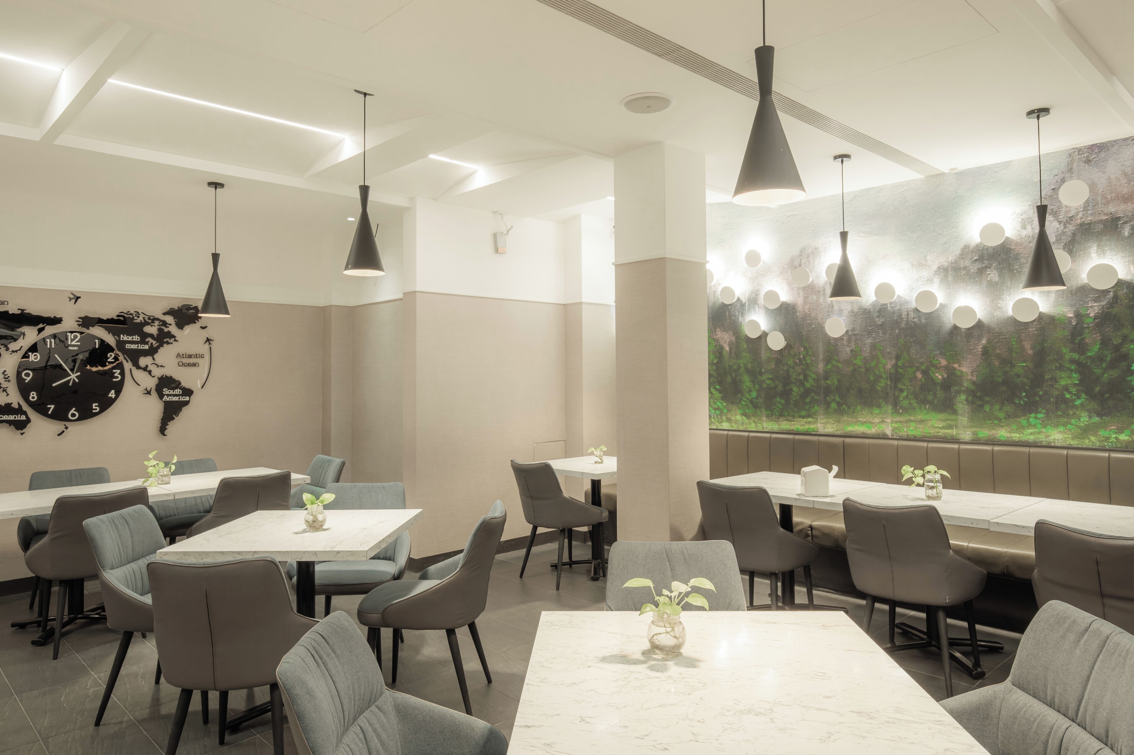 A modern dining area with round tables, grey upholstered chairs, pendant lighting, a world map on the wall, and a large window with a forest view.