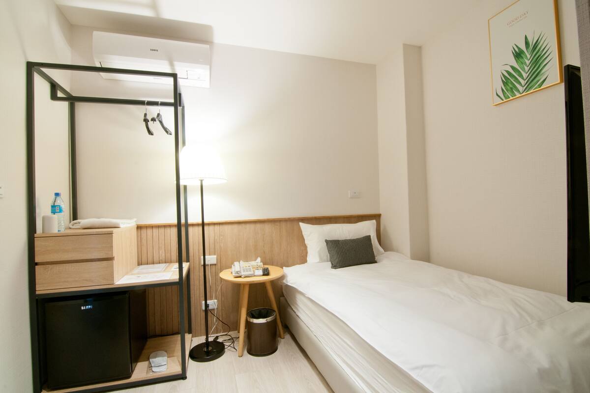 A hotel room with a bed, a bedside table with a lamp, a small desk with a phone, a mirror, and a trash bin.