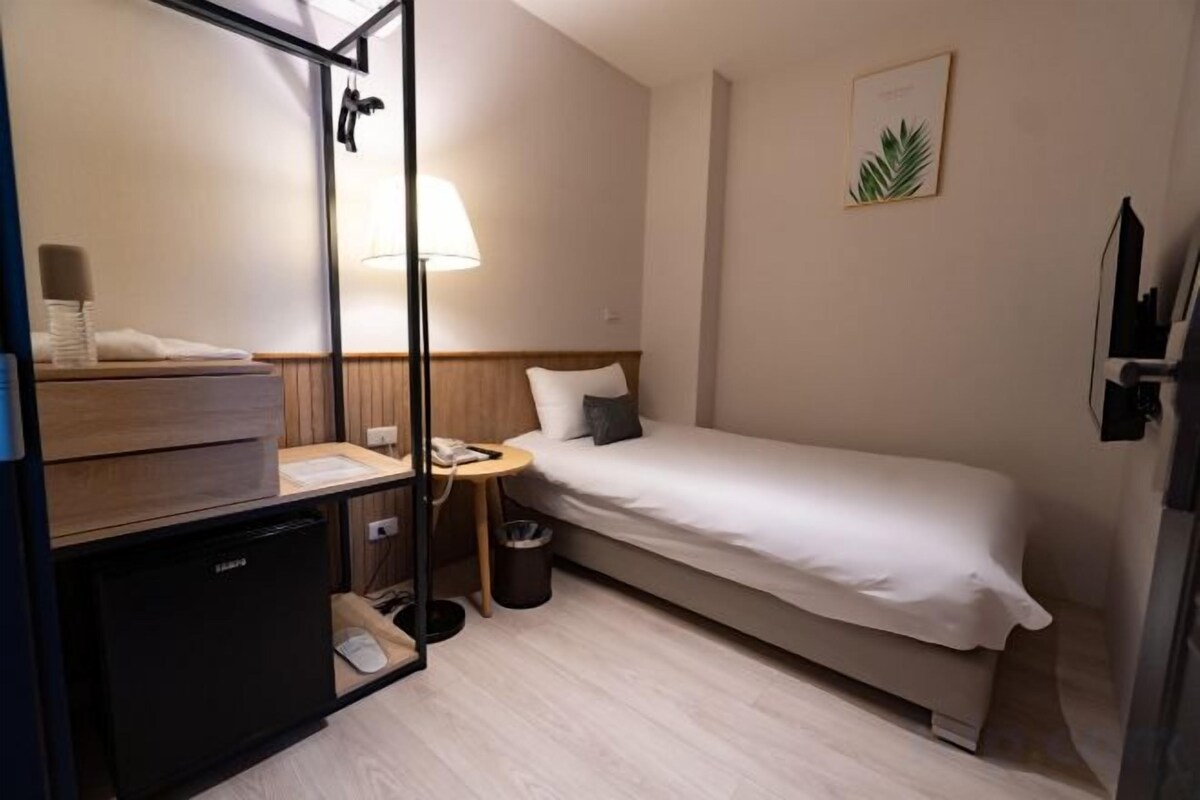 A compact hotel room with a single bed, a small desk, a lamp, a trash bin, and a television.
