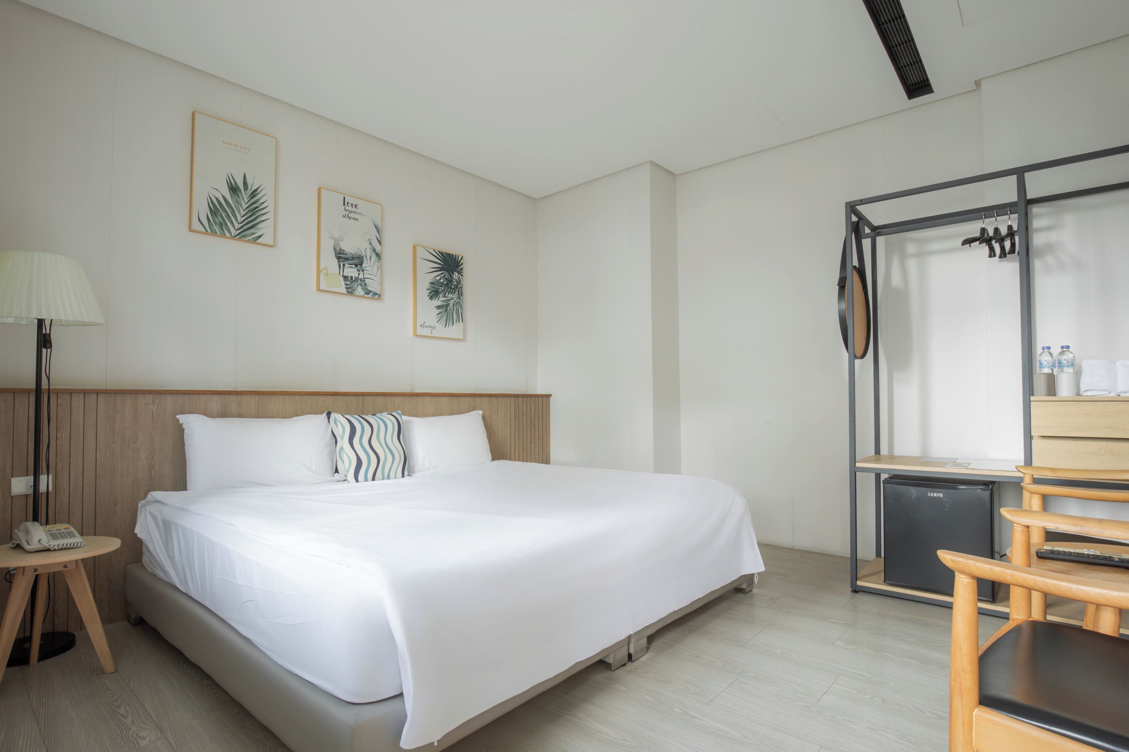 A hotel room with a bed, a desk with a lamp, a refrigerator, and wall art.