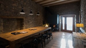 Un interno rustico con un lungo tavolo in legno, sedie nere, pareti in pietra, travi a vista e un camino.