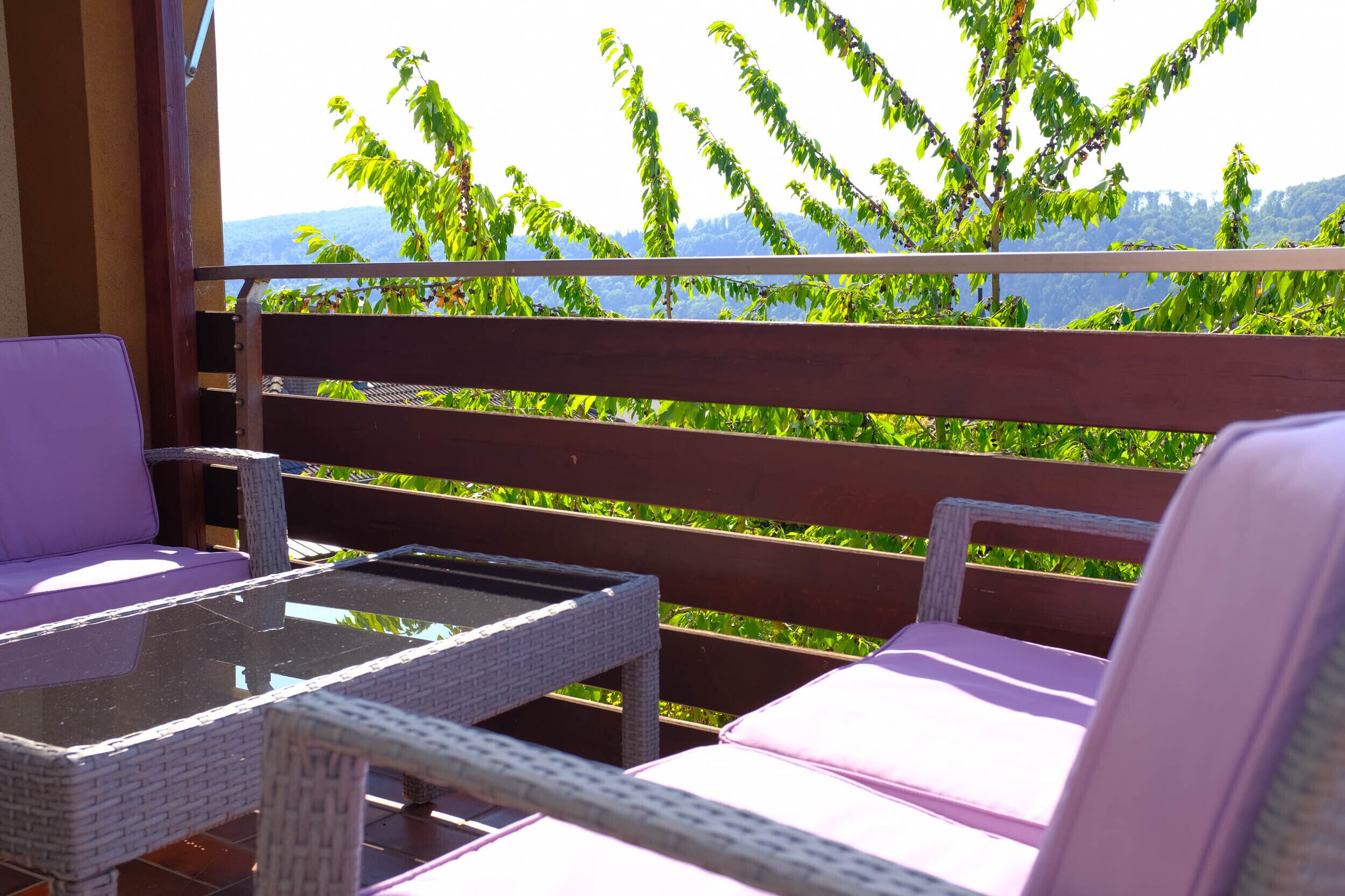 A balcony with wicker furniture and a glass table, overlooking a lush green landscape with trees and distant hills.