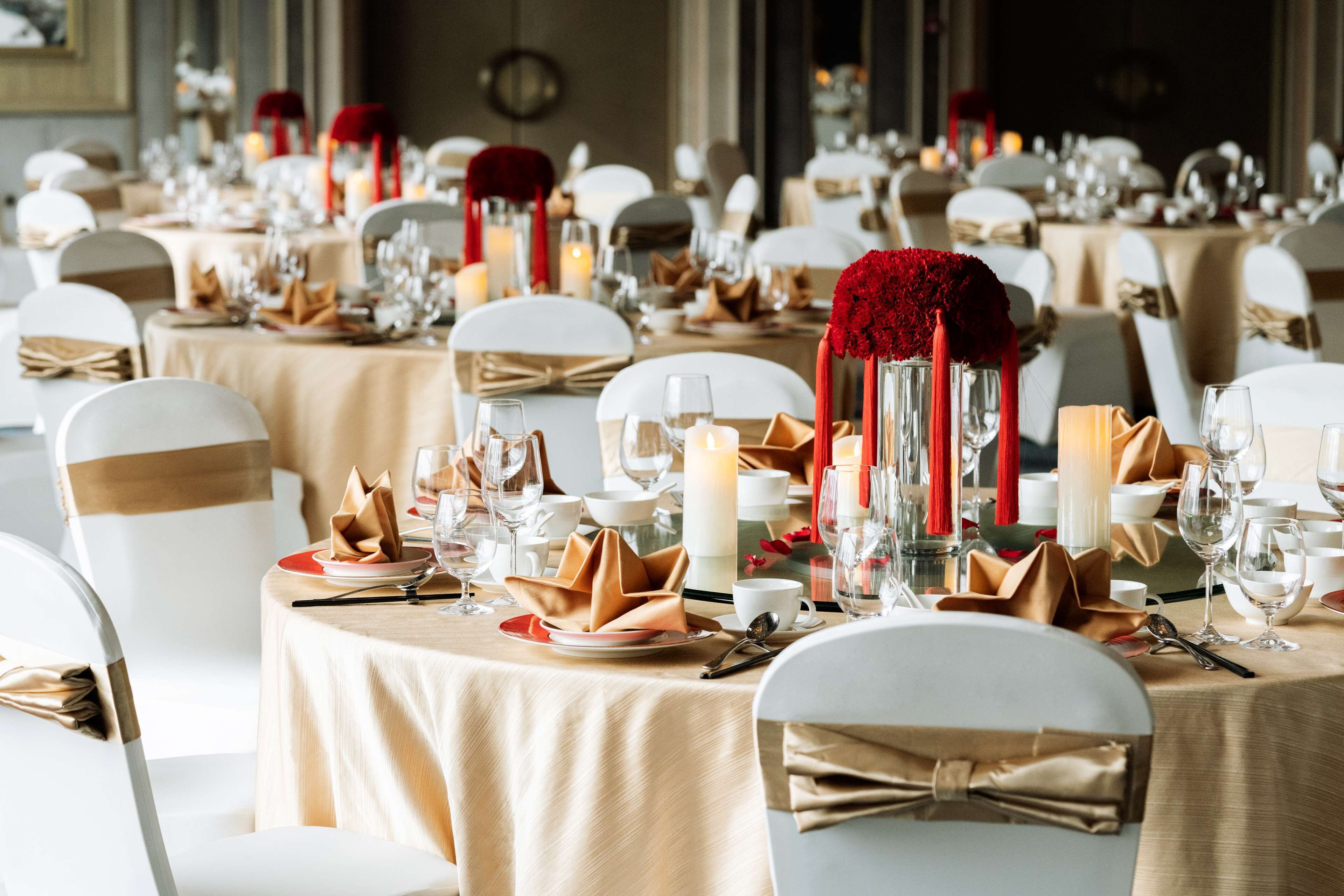 A banquet hall with round tables, white chairs, and red and white floral centerpieces.