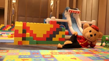 A play area with colorful building blocks, a teddy bear in a red uniform, and a slide.