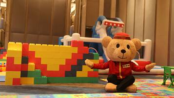 Un ours en peluche portant une veste rouge, assis sur un tapis de jeu coloré, avec une construction en LEGO en arrière-plan.