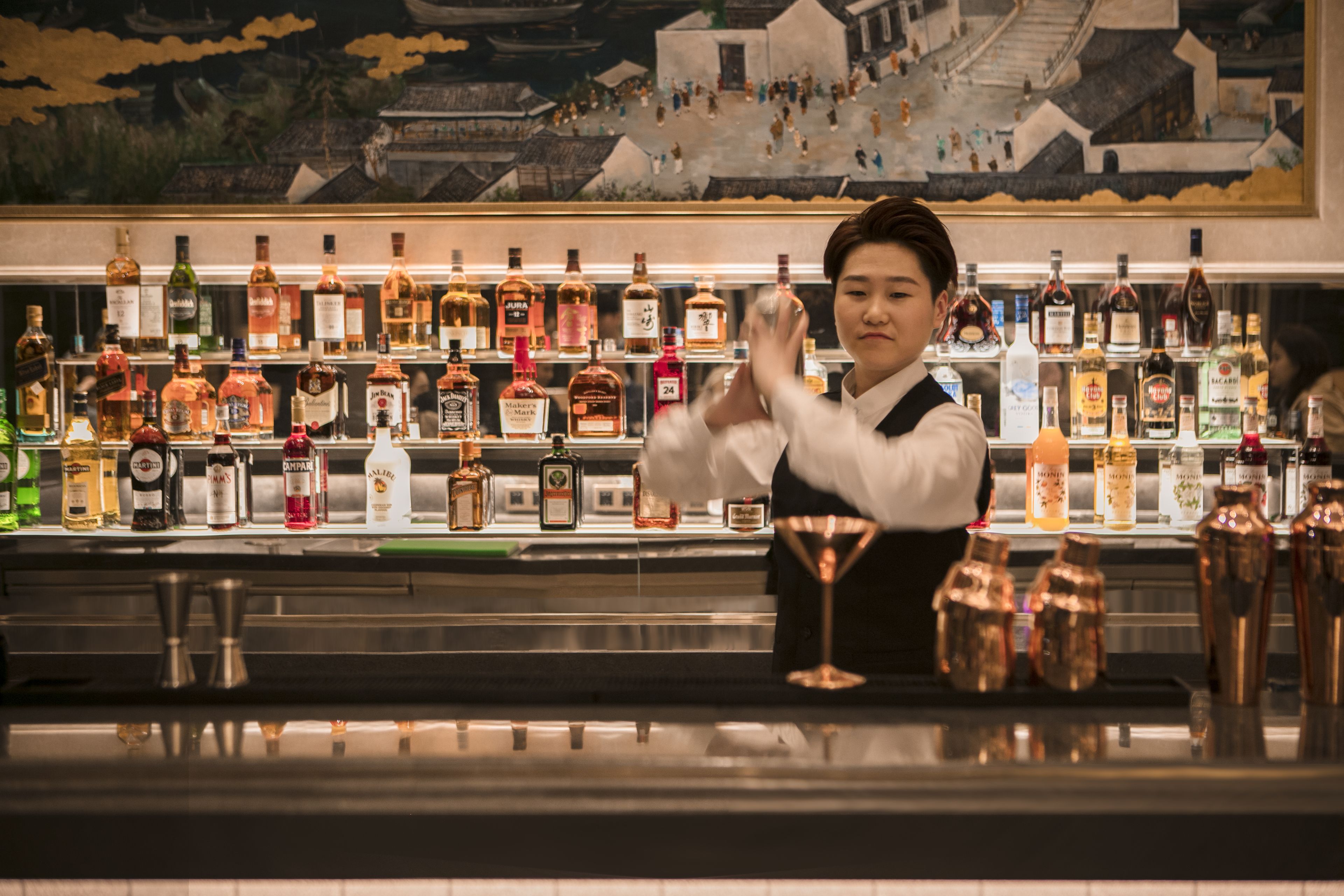 A bar with a bartender, shelves of liquor bottles, and a mural in the background.