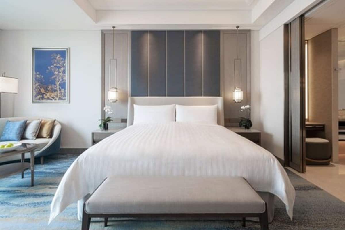 A modern hotel room with a large bed, a bench, a sofa, and a desk with a lamp.