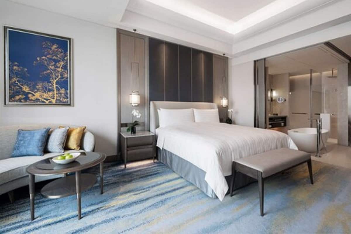 A modern hotel room with a large bed, a sitting area with a sofa and coffee table, and a bathroom with a bathtub and shower.