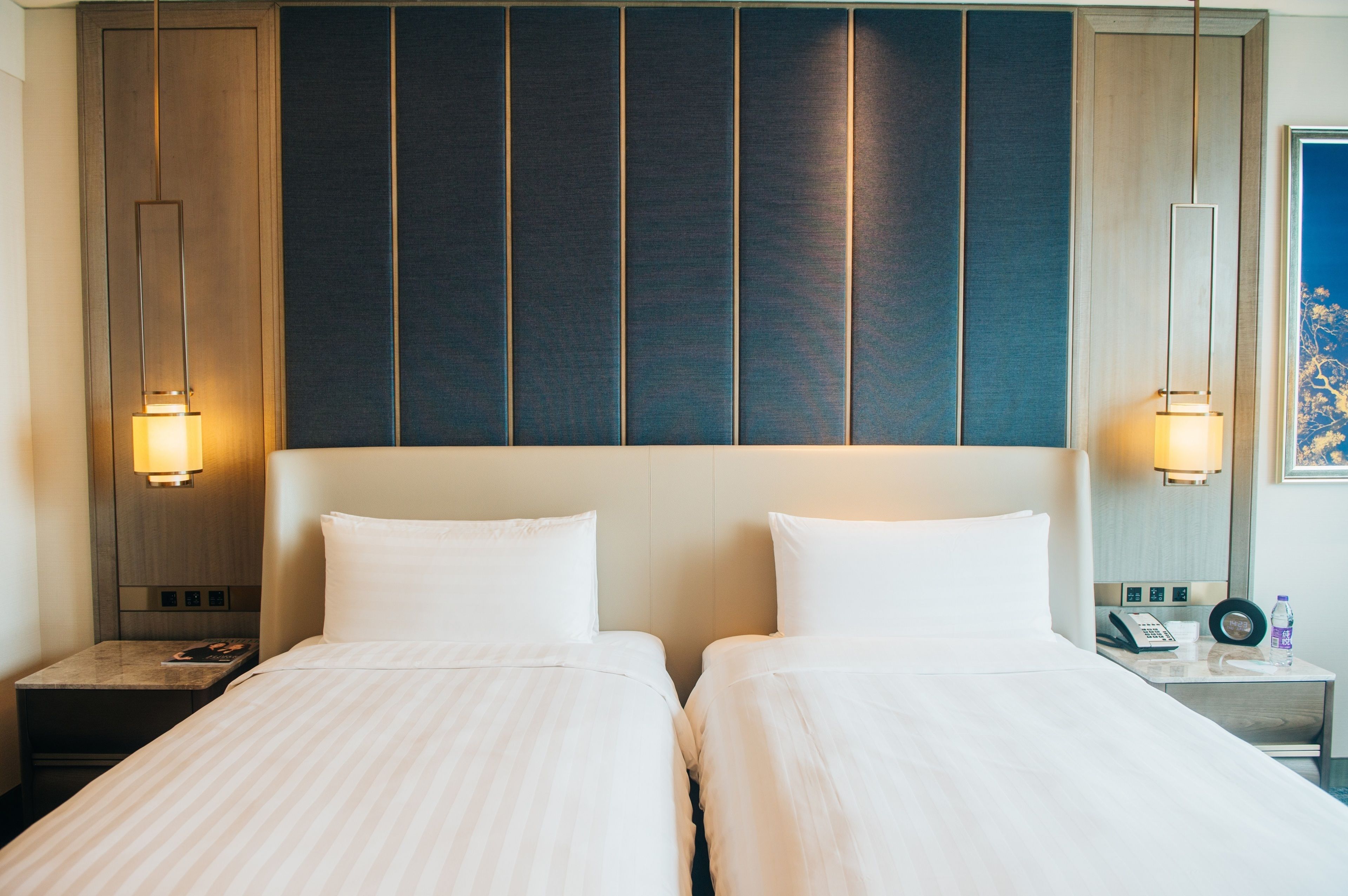 Ein Hotelzimmer mit zwei Betten, einem Holz-Kopfteil, Nachttischlampen, einem Nachttisch mit Telefon und einem gerahmten Bild an der Wand.