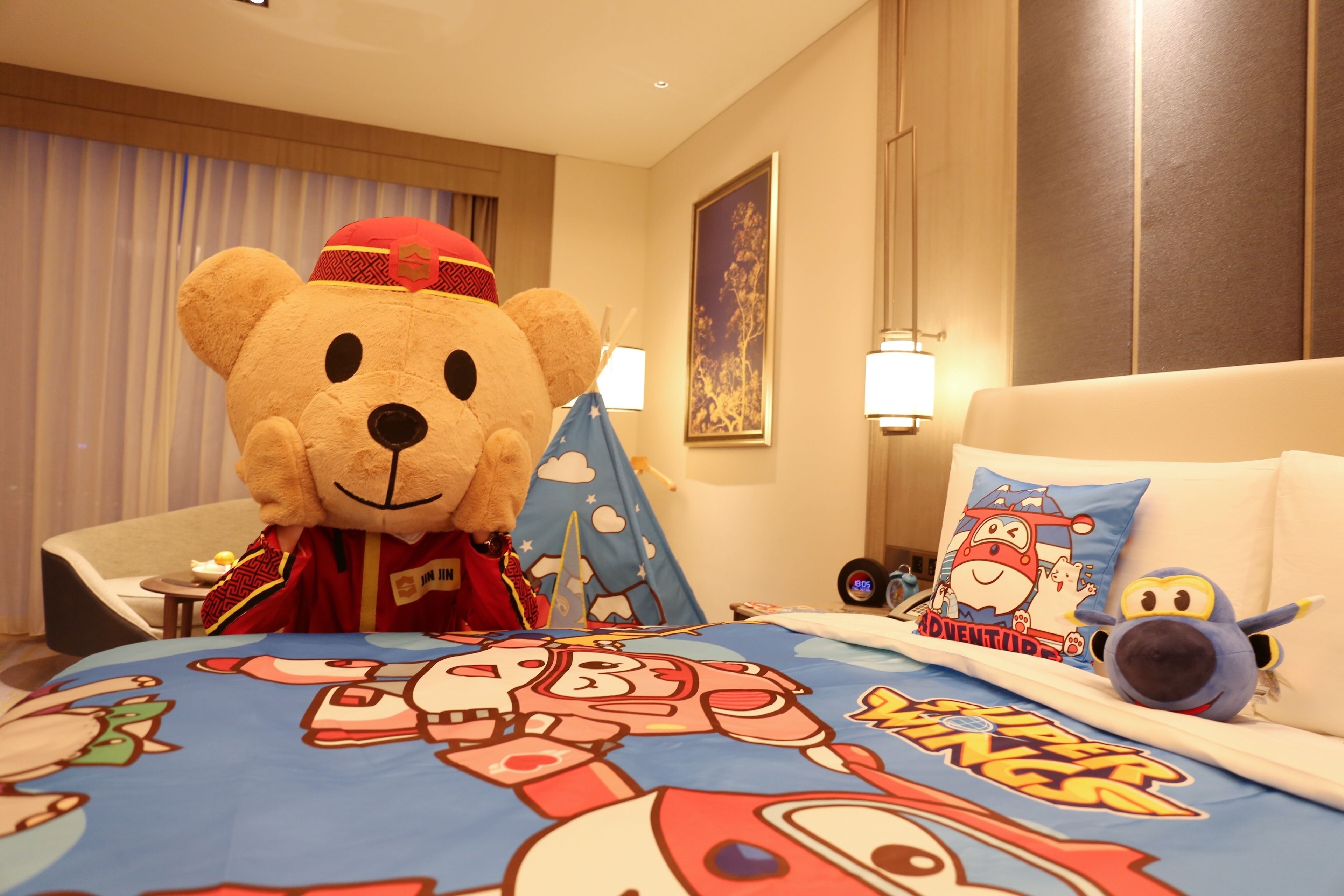Hotelkamer met een grote knuffelbeer, een bed met een kleurrijk dekbed met superheldenprint, een kussen met een cartoonfiguur, en een tafeltje met een lamp en een klok.