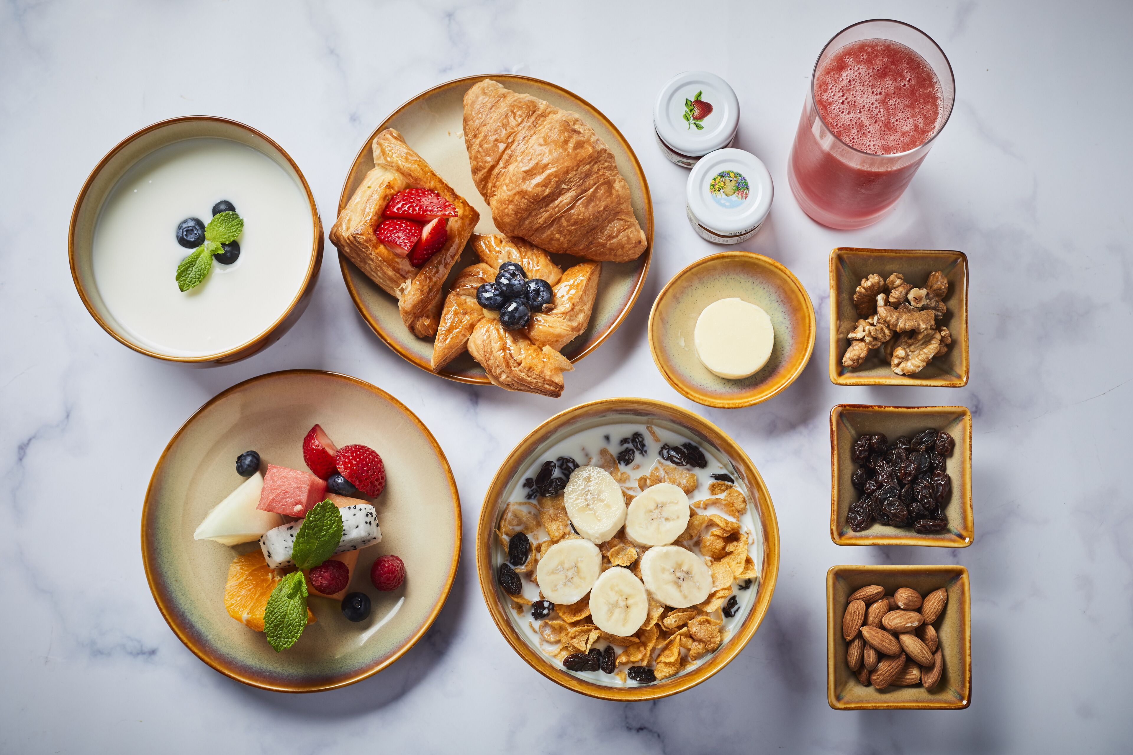 Variedad de alimentos para el desayuno, como yogur, bollería, frutas y frutos secos, sobre una superficie de mármol.