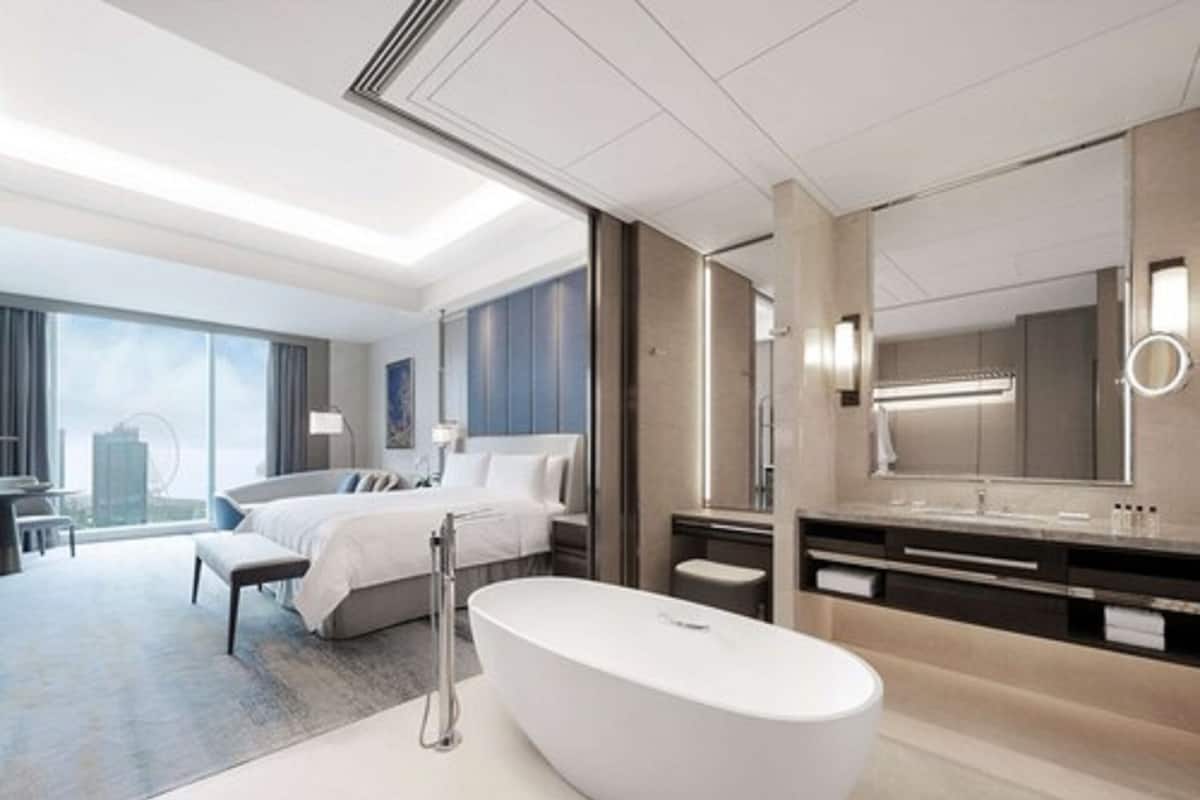 A modern hotel room with a large bed, a sitting area, a bathroom with a large mirror and bathtub, and a city view.
