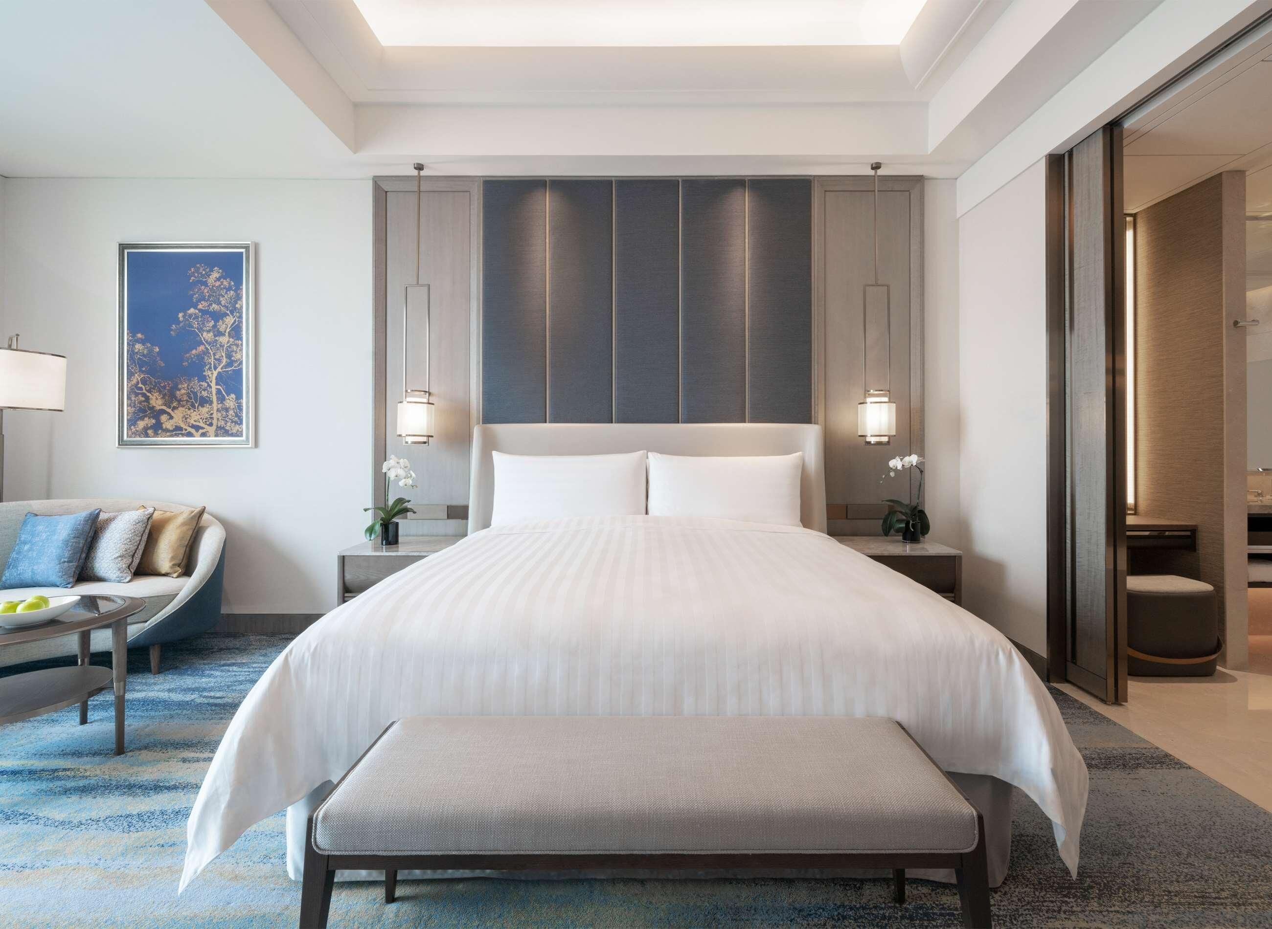 A modern hotel room with a large bed, bedside tables, a bench, a sofa, and a large window.