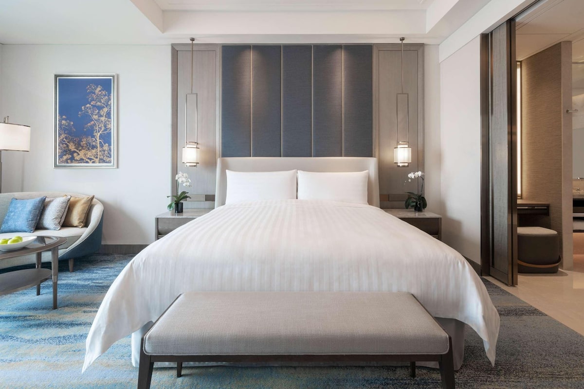 A modern hotel room with a large bed, bedside tables, a bench, a sofa, and a large window.