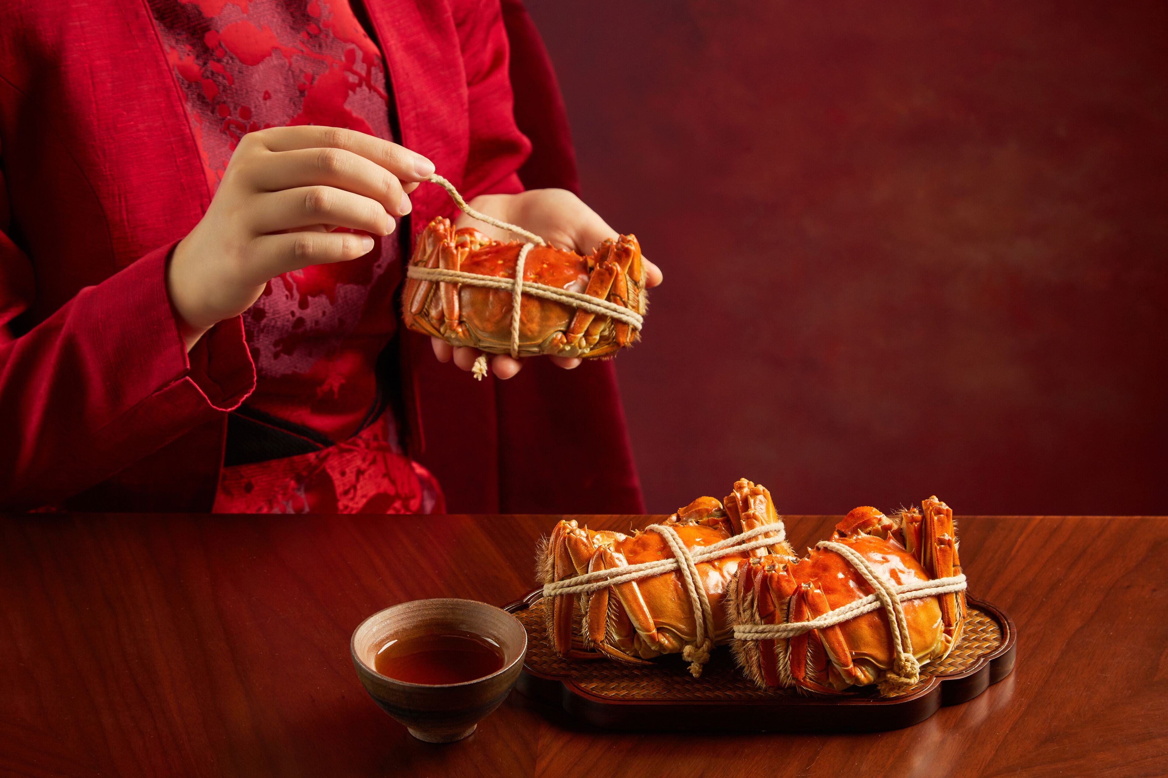 Une personne tenant un crabe attaché avec de la ficelle, avec un bol de sauce et un autre crabe sur un plateau.