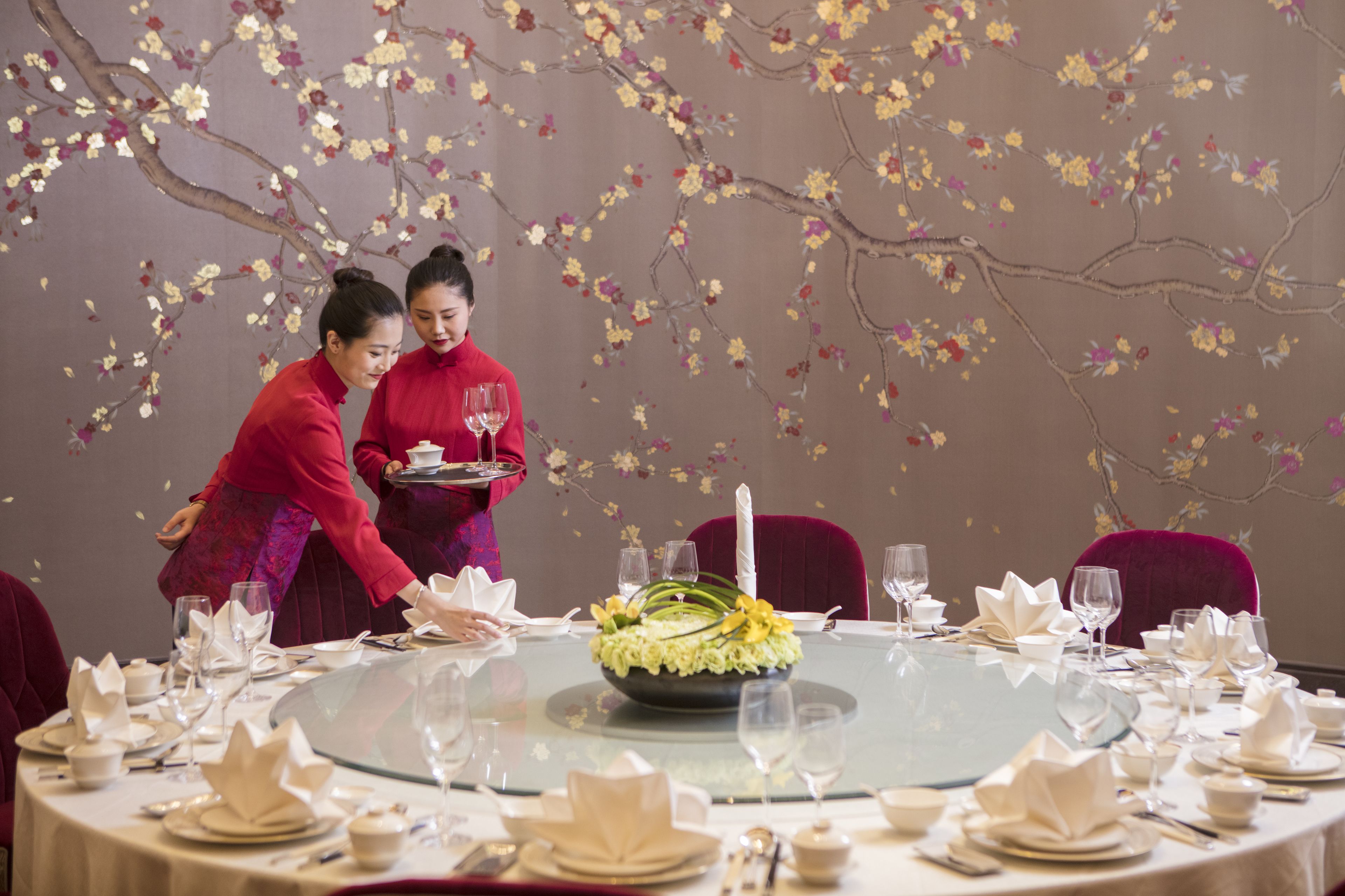 A round dining table set for a meal with a floral centerpiece, surrounded by chairs and a decorative wall.
