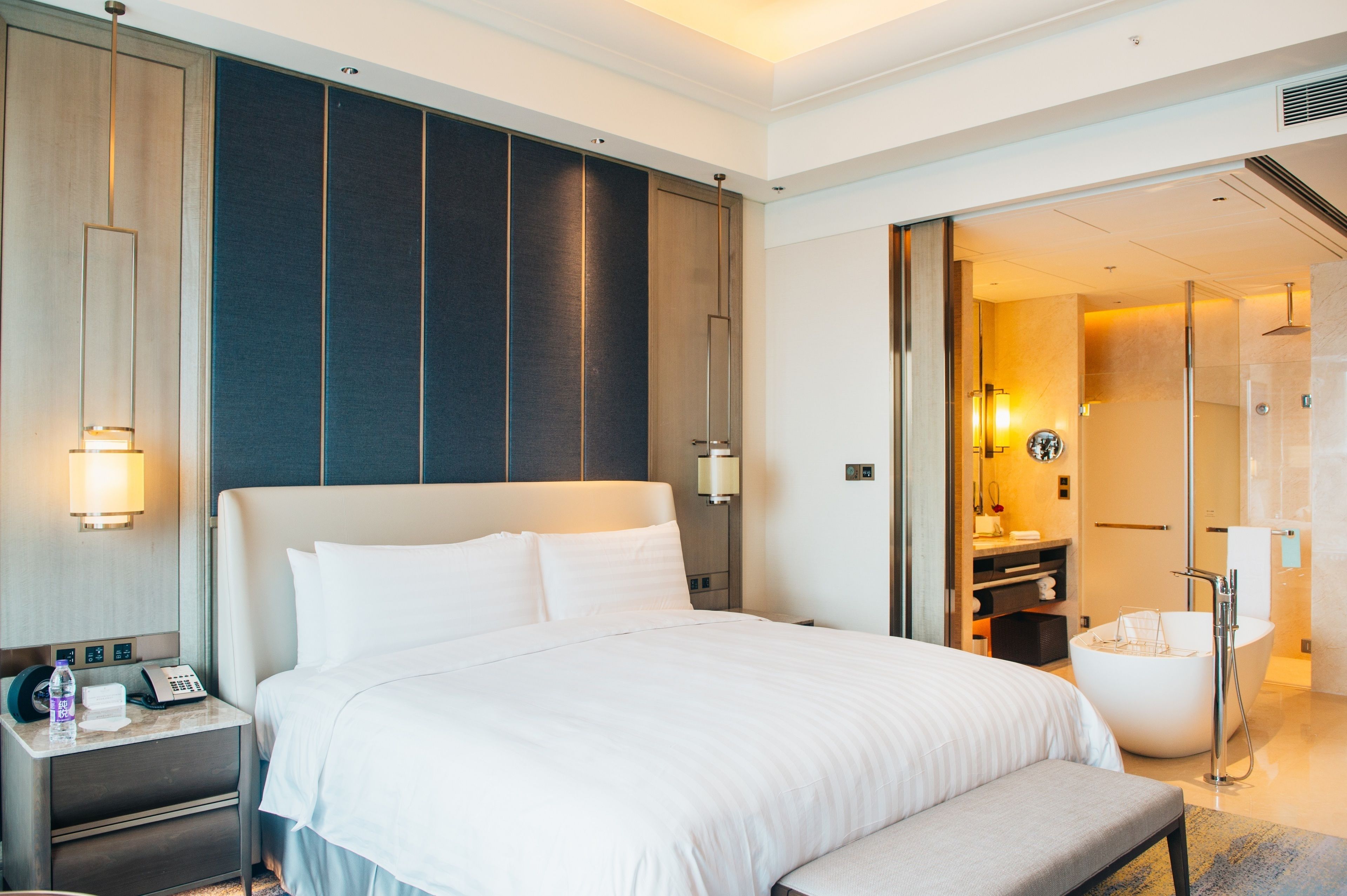 Ein modernes Hotelzimmer mit einem großen Bett, einem Nachttisch mit Telefon und Wasserflasche, einem Badezimmer mit Badewanne und einem begehbaren Kleiderschrank.