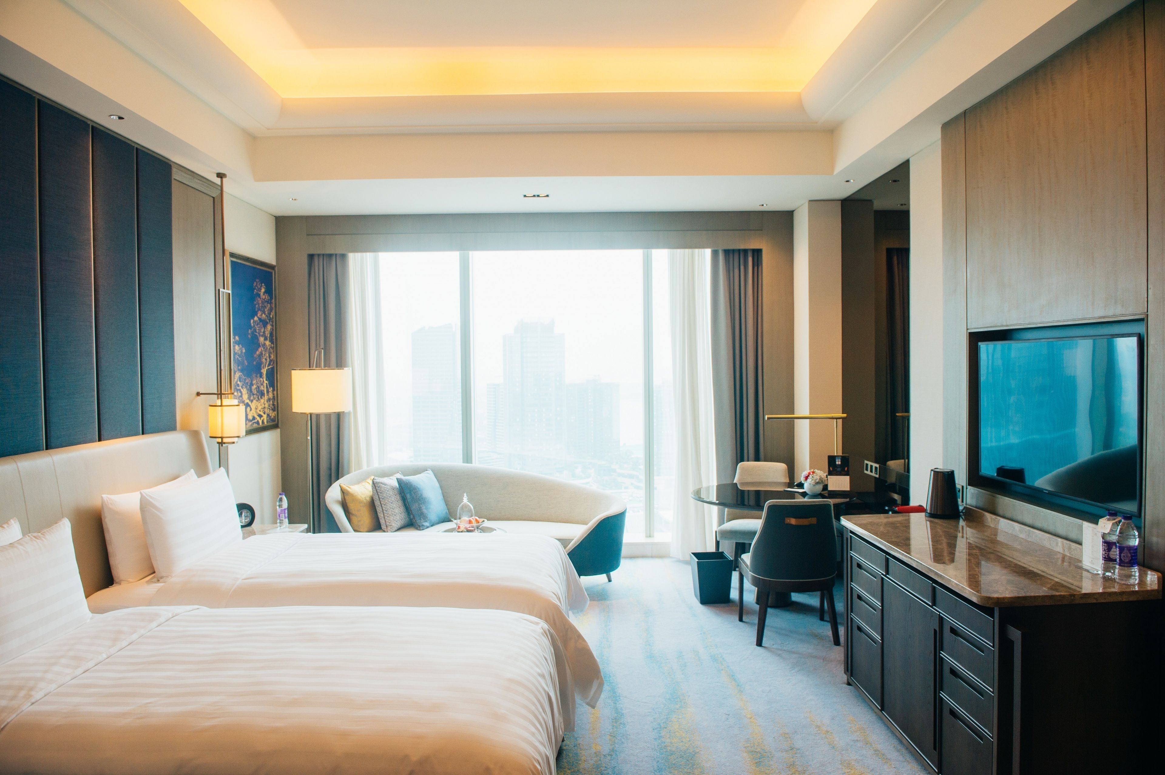 Een hotelkamer met een groot bed, een bureau met stoel, een televisie en uitzicht op de stad door grote ramen.