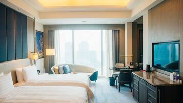 Een hotelkamer met een groot bed, een bureau met stoel, een televisie en uitzicht op de stad door grote ramen.