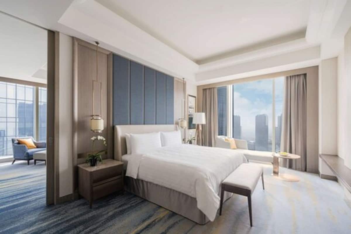 A modern hotel room with a large bed, a desk, and a chair. There is a large window with a city view.