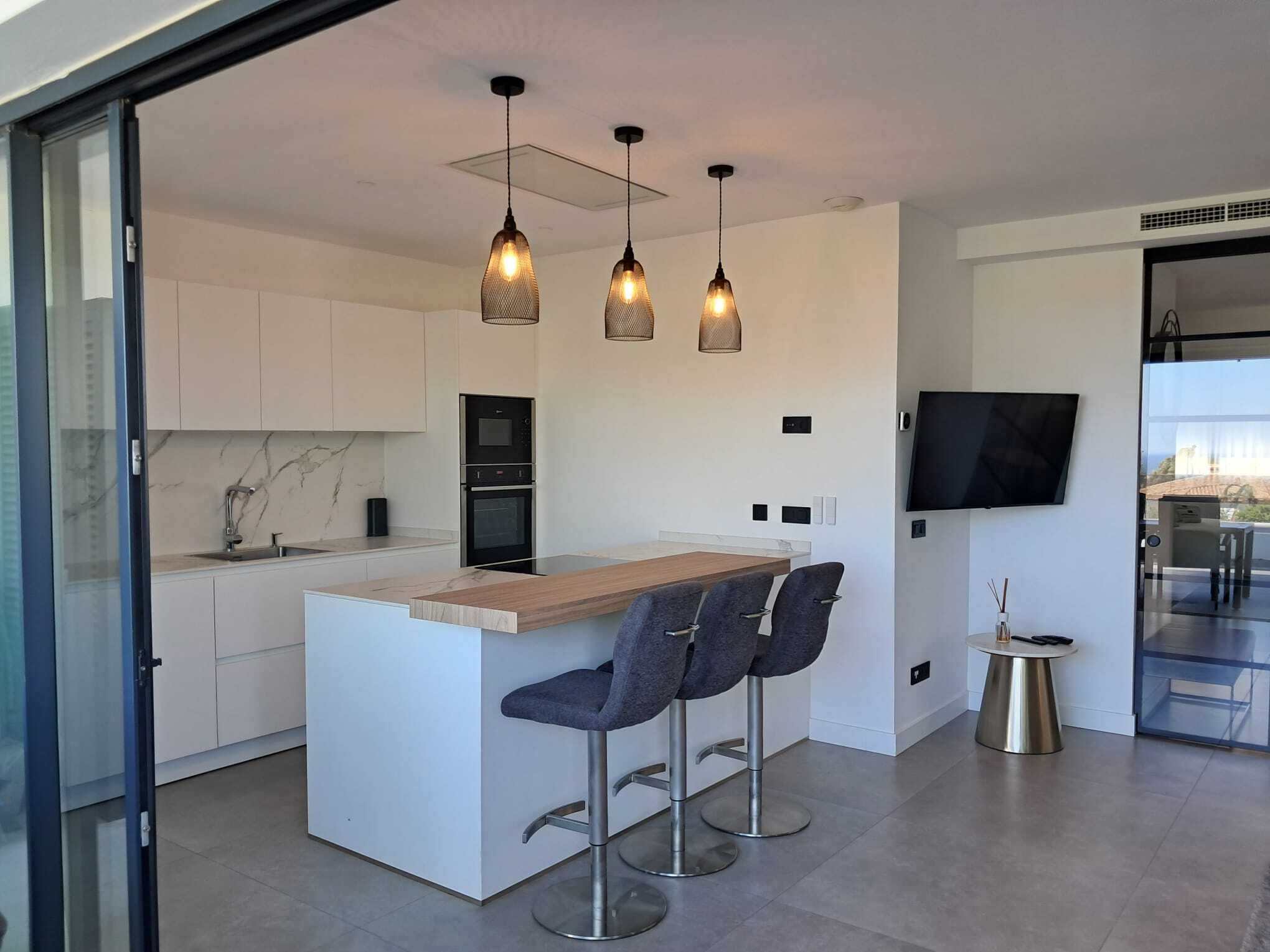 A modern kitchen with a central island, bar stools, pendant lighting, and a flat-screen TV mounted on the wall.