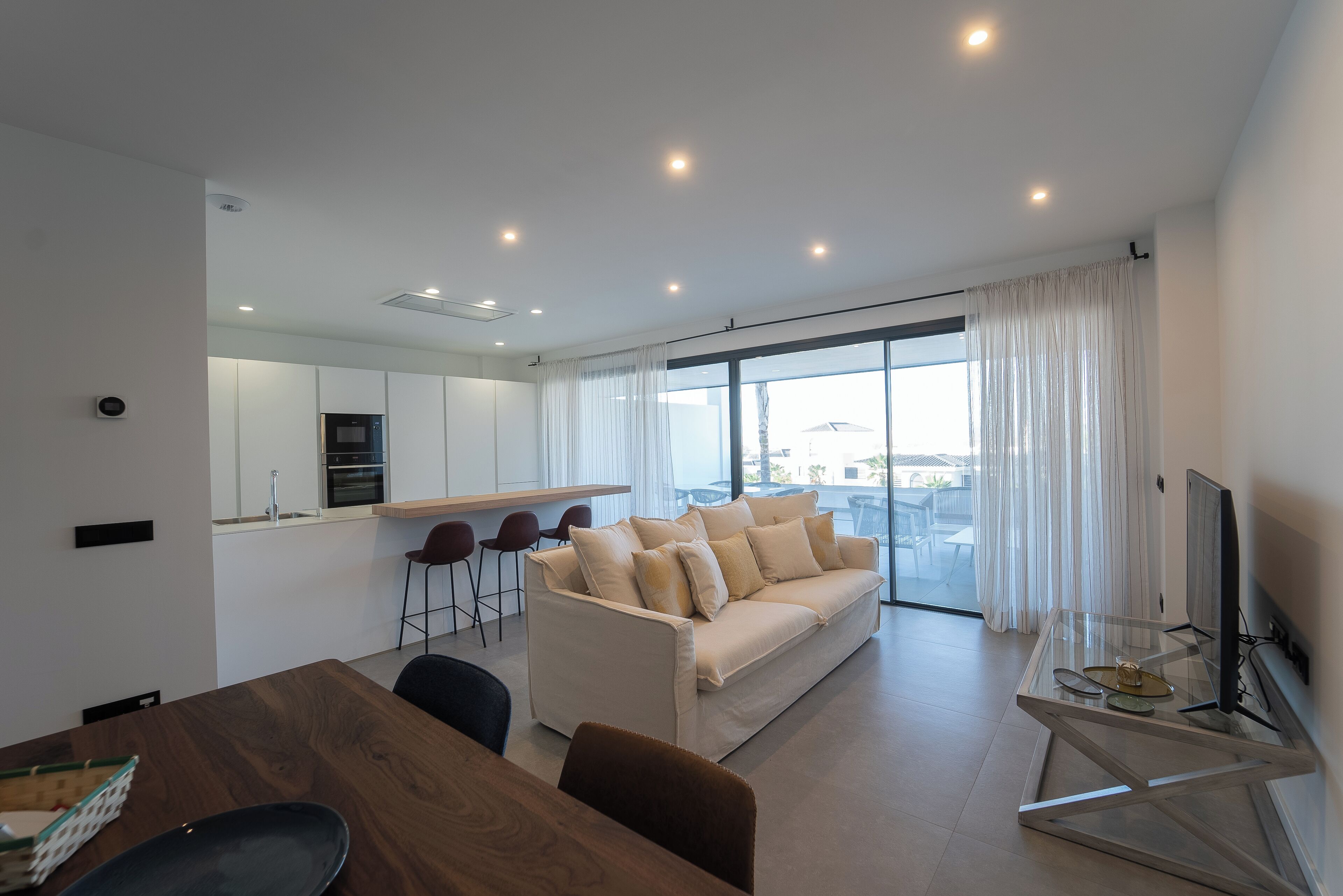 Una sala de estar moderna con un sofá, una mesa de centro de vidrio y un televisor. Cuenta con una zona de cocina con un refrigerador y una mesa de comedor con sillas.
