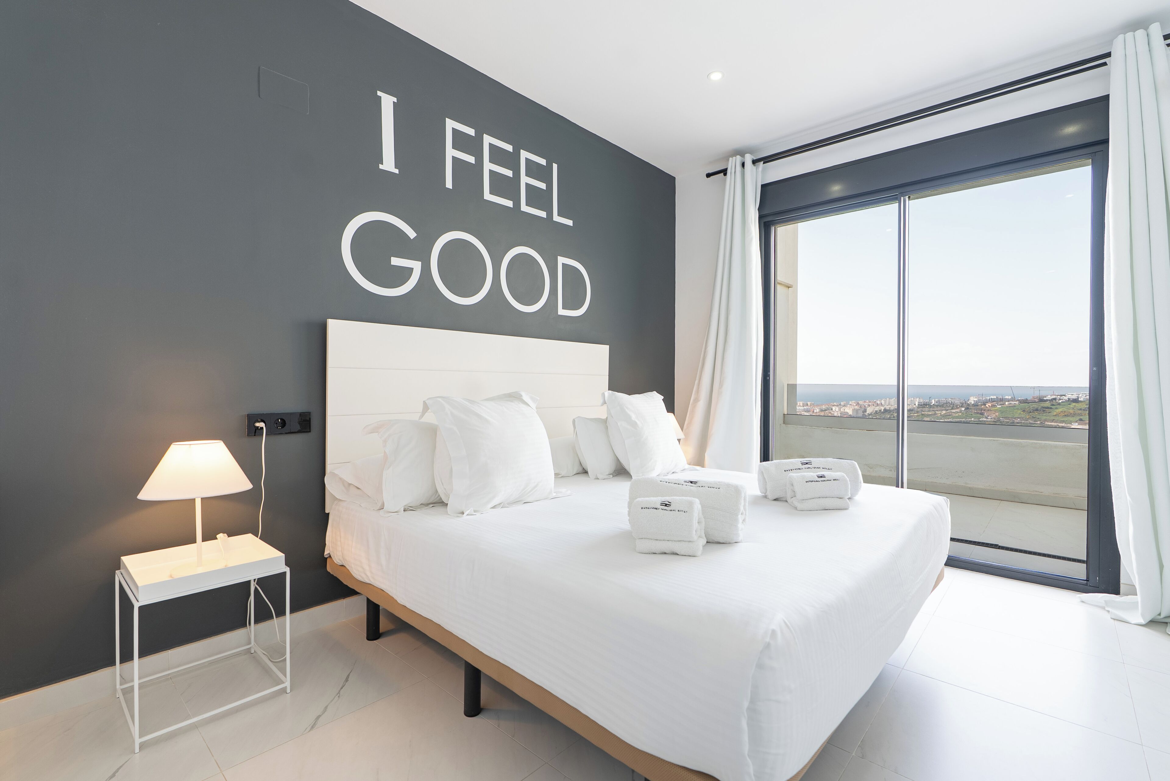 Une chambre d’hôtel moderne avec un grand lit, une table de chevet avec une lampe, une vue sur la ville grâce à de grandes fenêtres et une citation inspirante affichée sur le mur.
