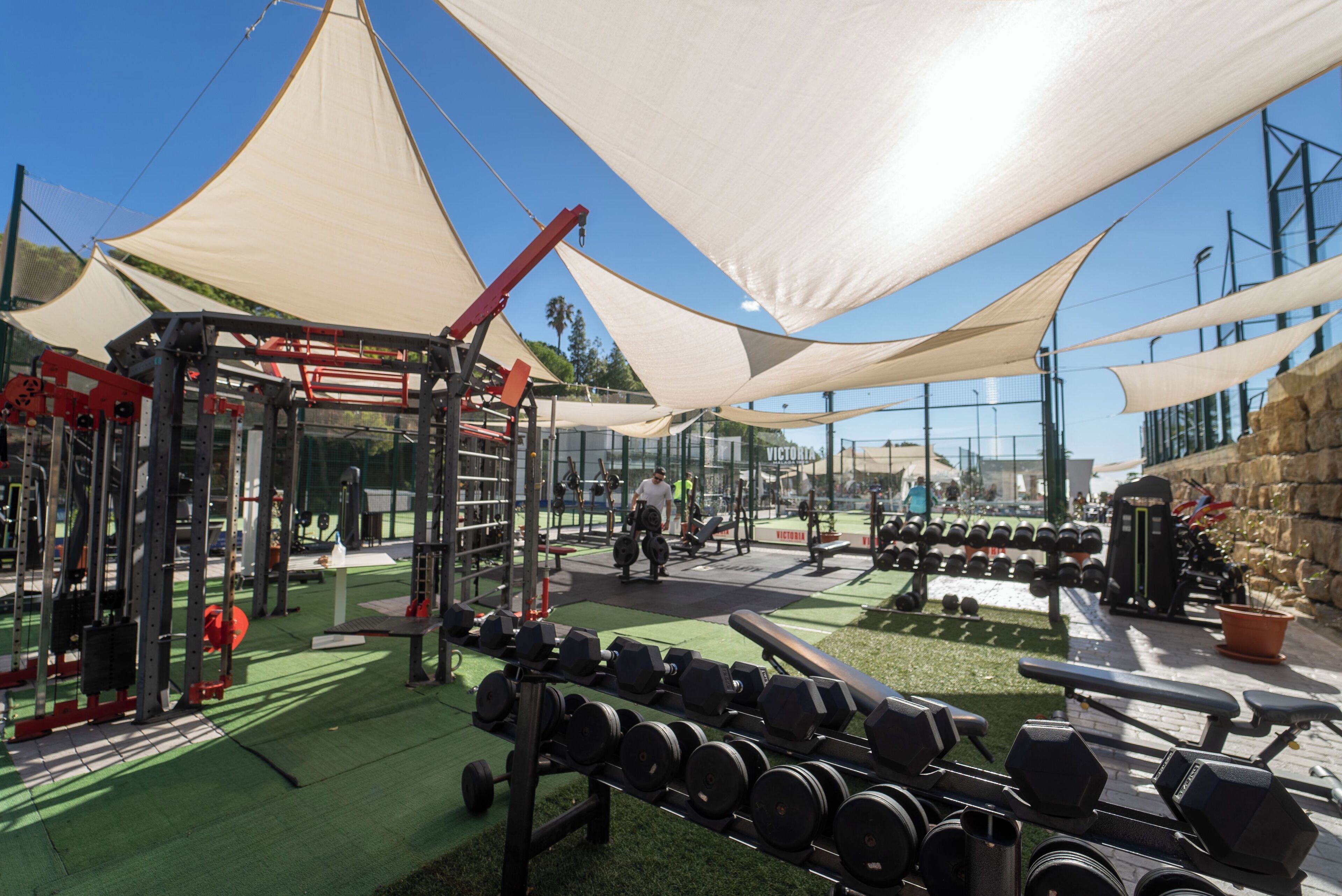 Un gimnasio con diverso equipo de ejercicio, incluyendo mancuernas y máquinas de pesas, bajo grandes estructuras de sombra.