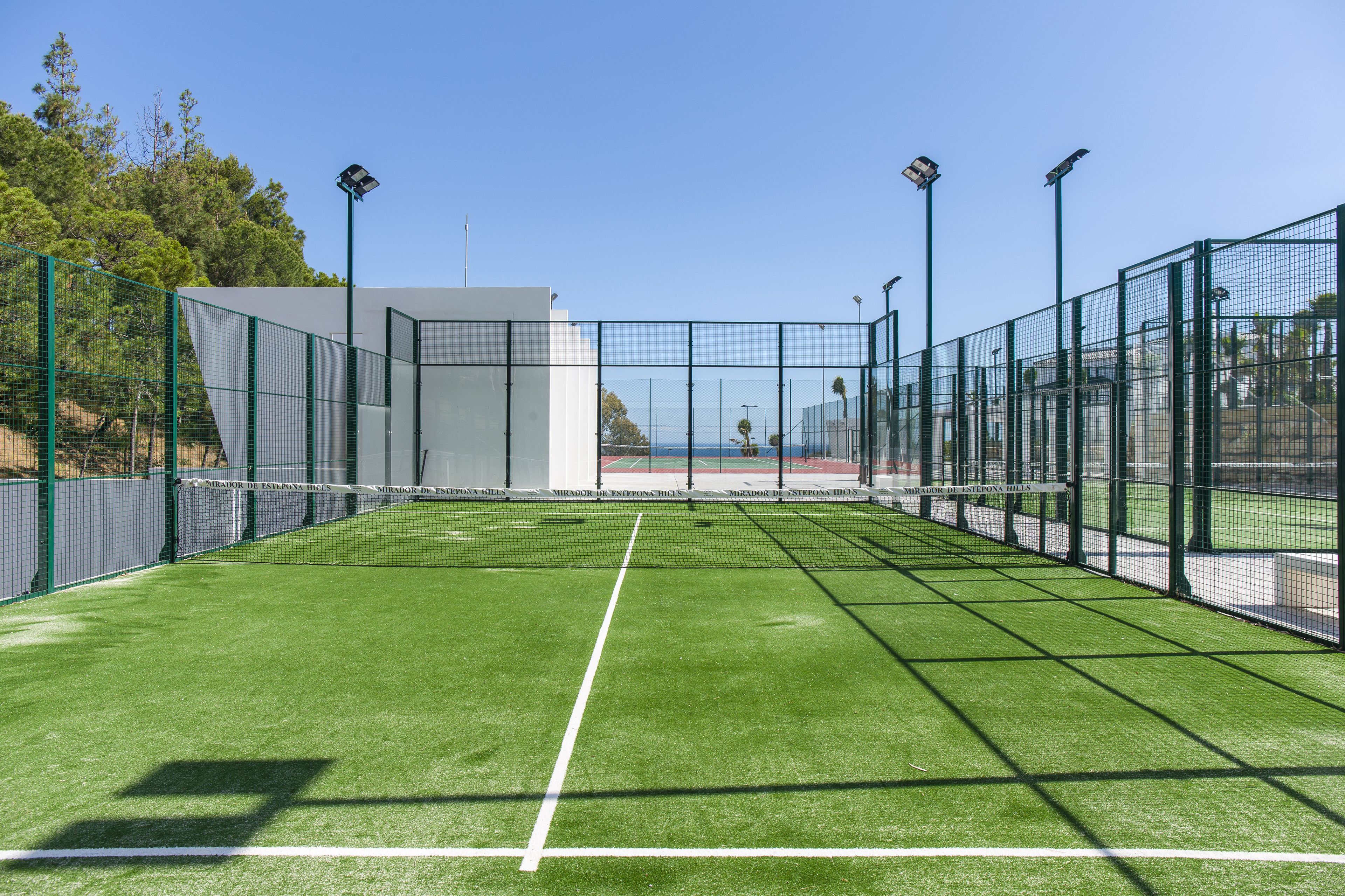 An indoor tennis court with a green surface, surrounded by a green fence, and equipped with overhead lighting.