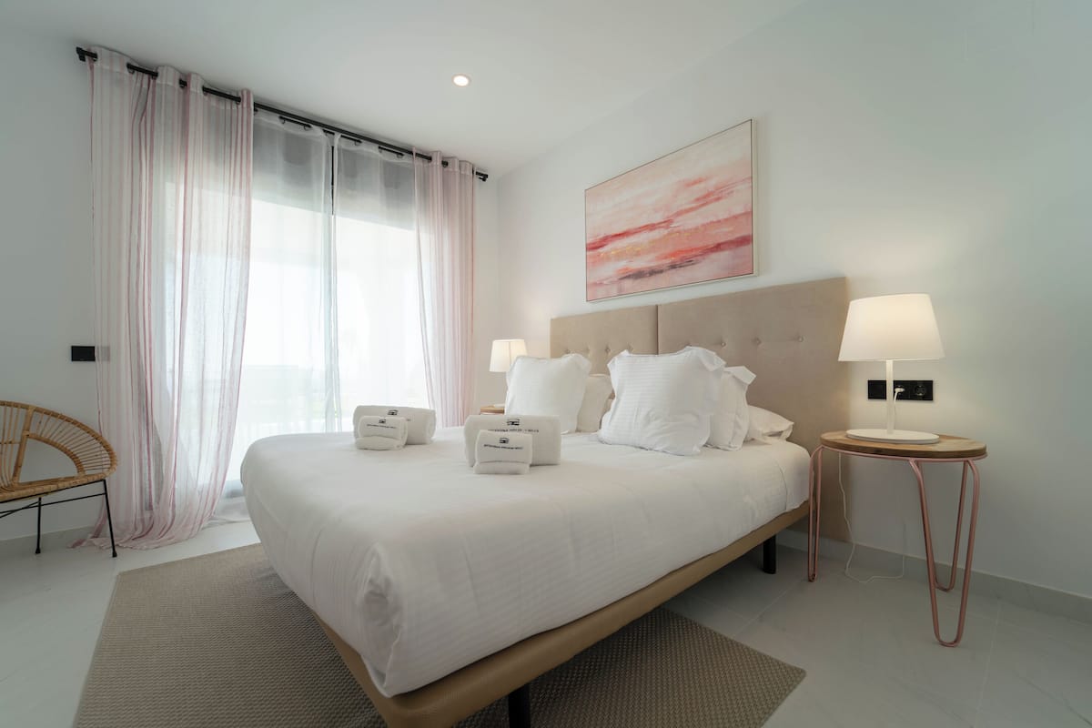 Une chambre d’hôtel moderne avec un grand lit, deux tables de chevet avec des lampes, une tête de lit, une œuvre d’art accrochée au mur et des rideaux légers.