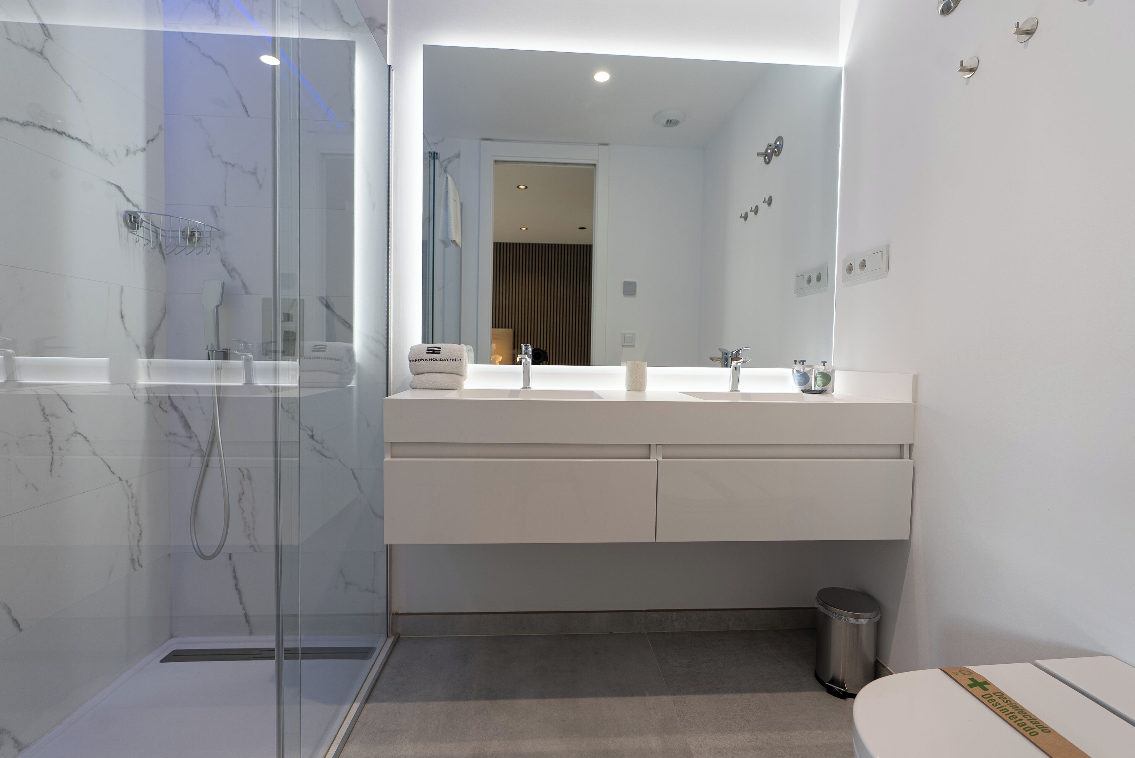 Un baño moderno con ducha envuelta en vidrio, un tocador blanco con dos lavamanos y un espejo grande.