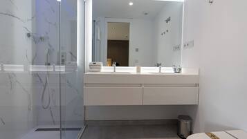 Un baño moderno con ducha envuelta en vidrio, un tocador blanco con dos lavamanos y un espejo grande.
