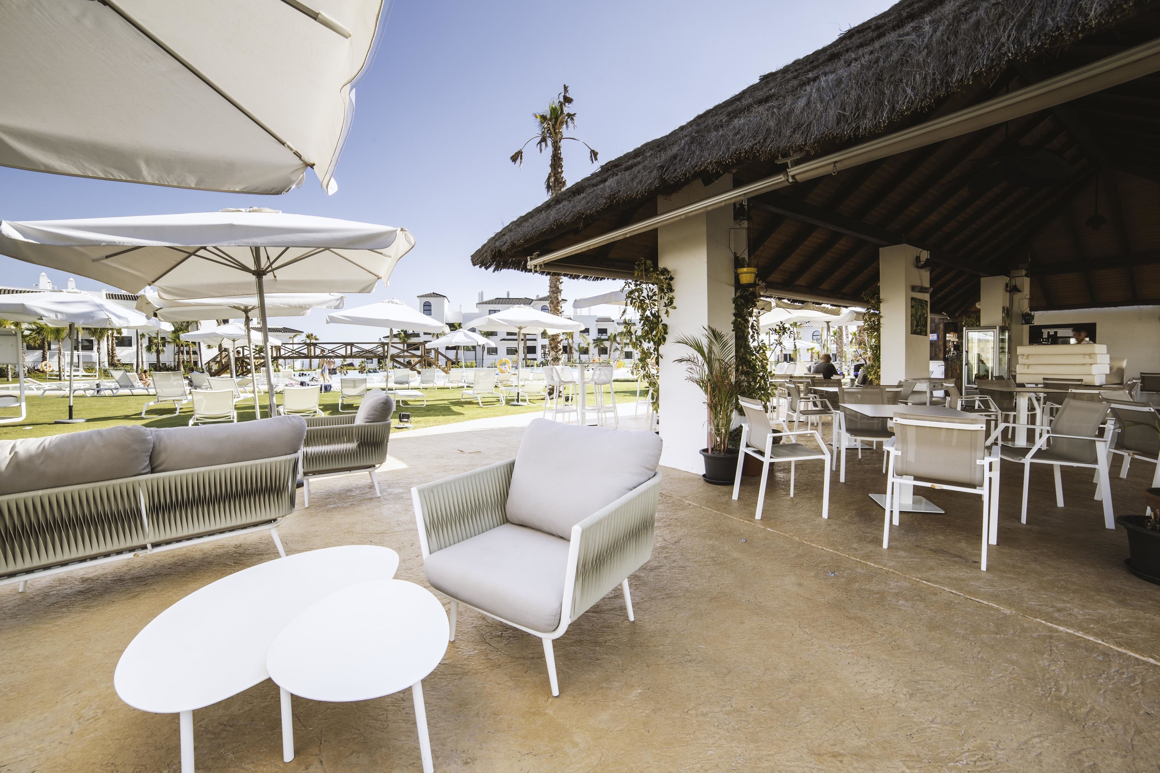 Un espace extérieur aménagé avec des meubles blancs et des parasols, une structure à toit de chaume et une piscine en arrière-plan.