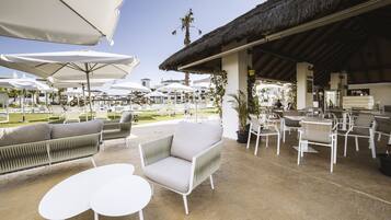 Un espace extérieur aménagé avec des meubles blancs et des parasols, une structure à toit de chaume et une piscine en arrière-plan.