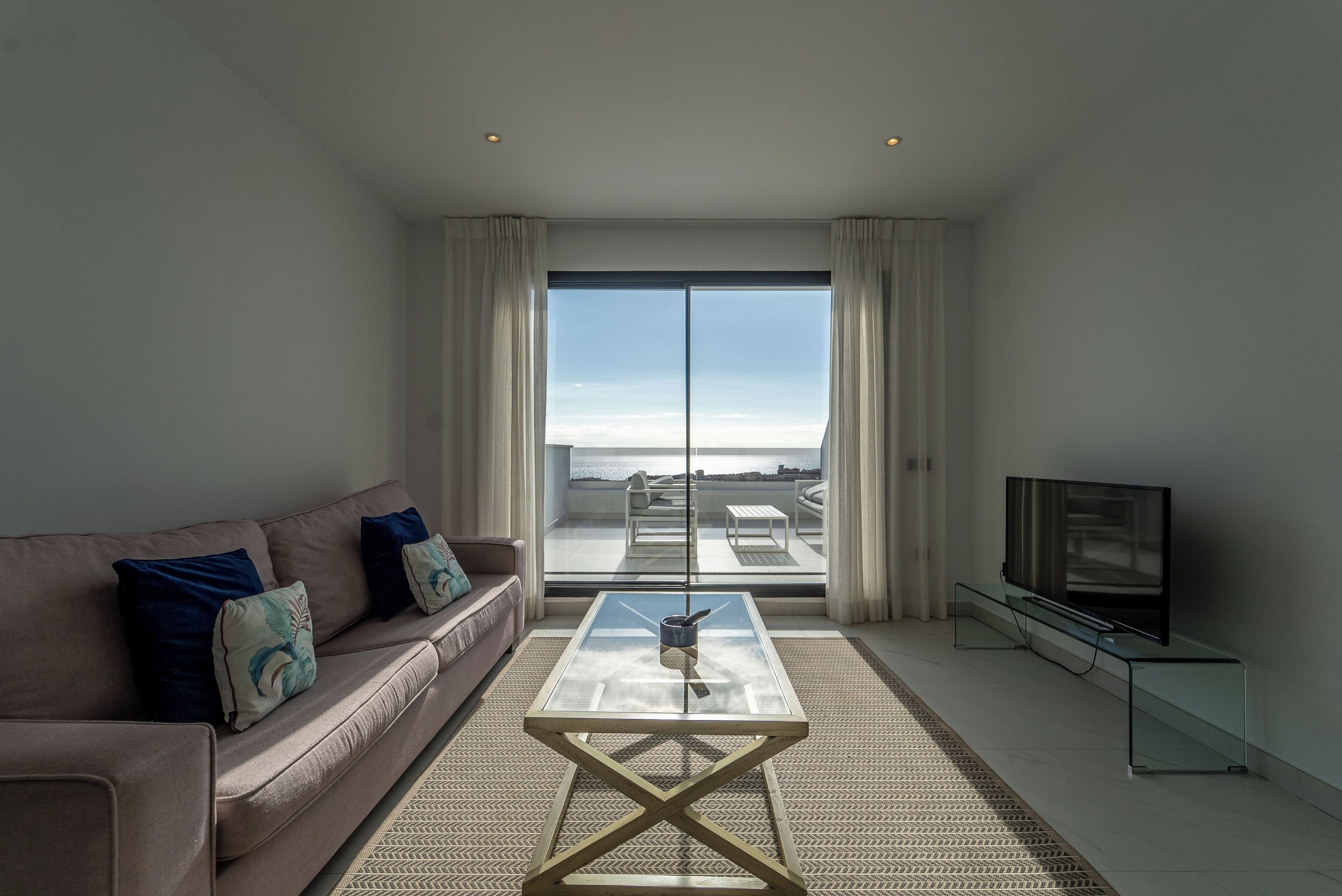 Una sala de estar moderna con un sofá, una mesa de centro de vidrio, un televisor sobre un mueble bajo y un amplio ventanal corredero que da a un balcón con vistas.