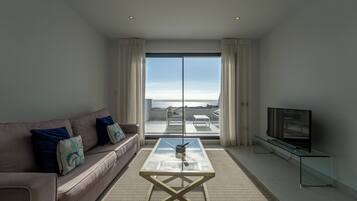 Una sala de estar moderna con un sofá, una mesa de centro de vidrio, un televisor sobre un mueble bajo y un amplio ventanal corredero que da a un balcón con vistas.