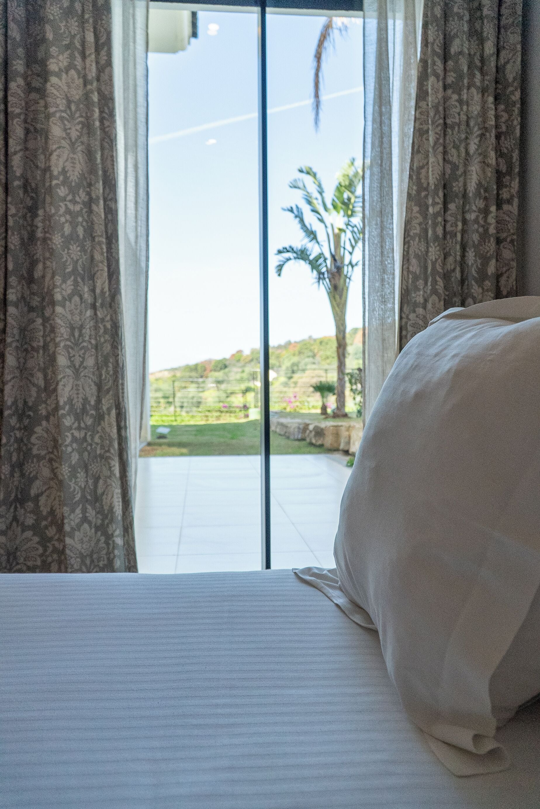 A bedroom with a large bed, a view of a garden through a sliding glass door, and curtains.